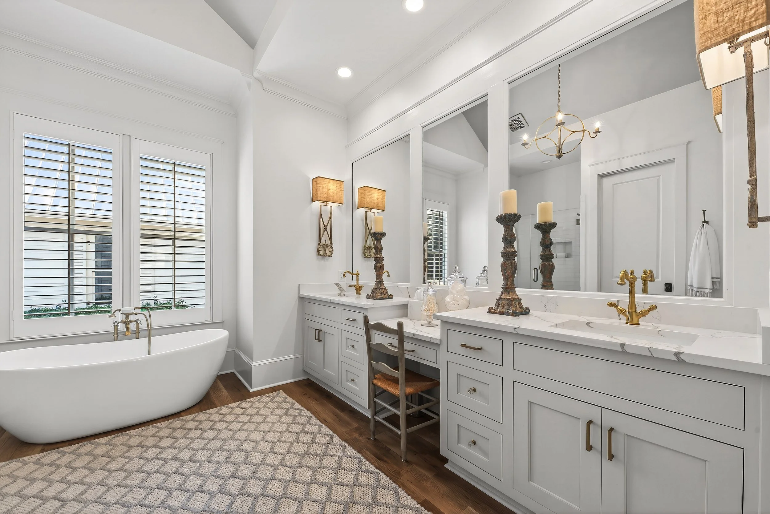 Bright bathroom with large window, white clawfoot tub, white cabinetry, gold fixtures, decorative candlesticks, and a brown and beige area rug.