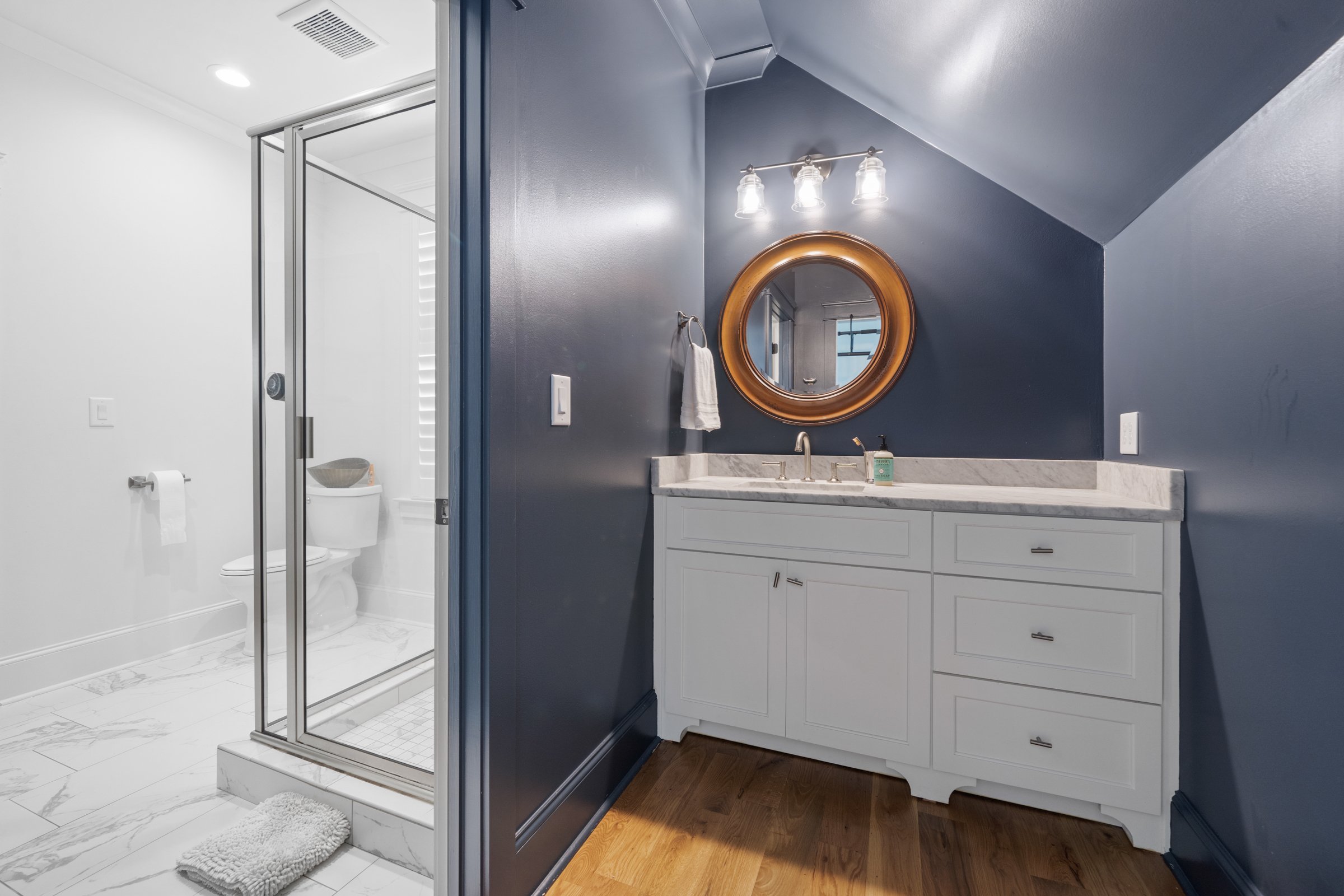 Bathroom with dark blue wall, white vanity with marble countertop, oval mirror, and three light fixtures above. Adjacent shower with glass door, white tiles, and a gray rug on the wooden floor.