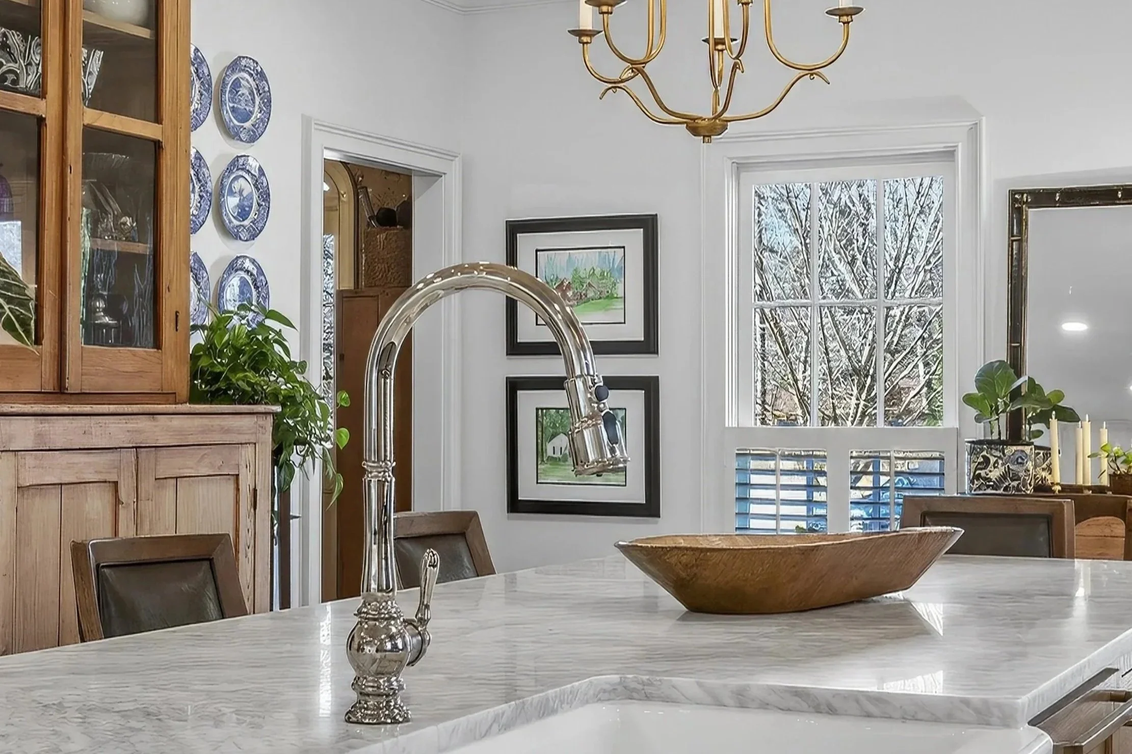 A bright kitchen with a white marble countertop island, a metallic faucet, wooden cabinets, and a window revealing snowy trees outside. There are framed pictures on the wall, a chandelier hanging from the ceiling, and various plants and decorative items.