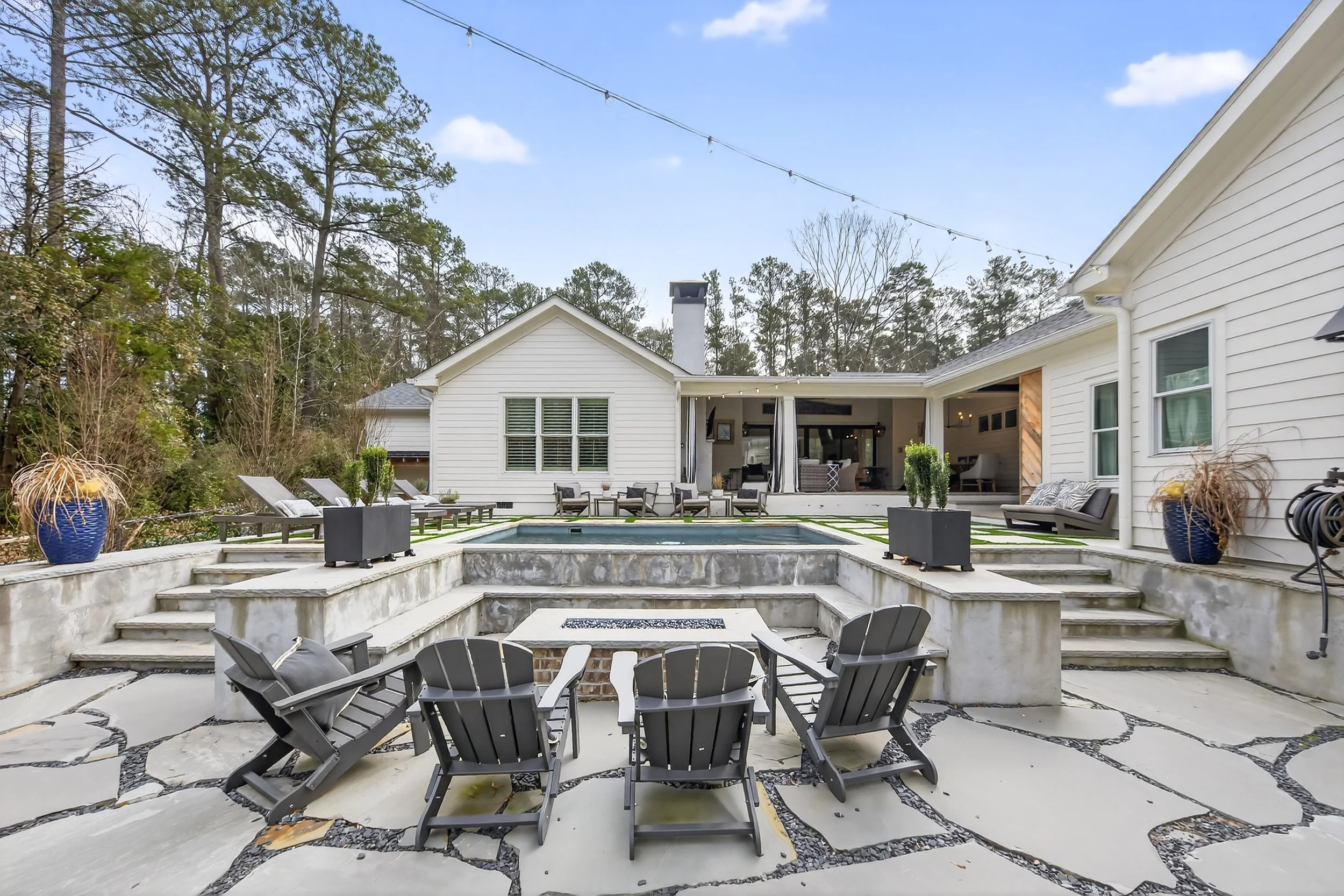 Wide view of a backyard with a pool, lounge chairs, outdoor seating, potted plants, and a white house with a deck, surrounded by trees under a partly cloudy sky.