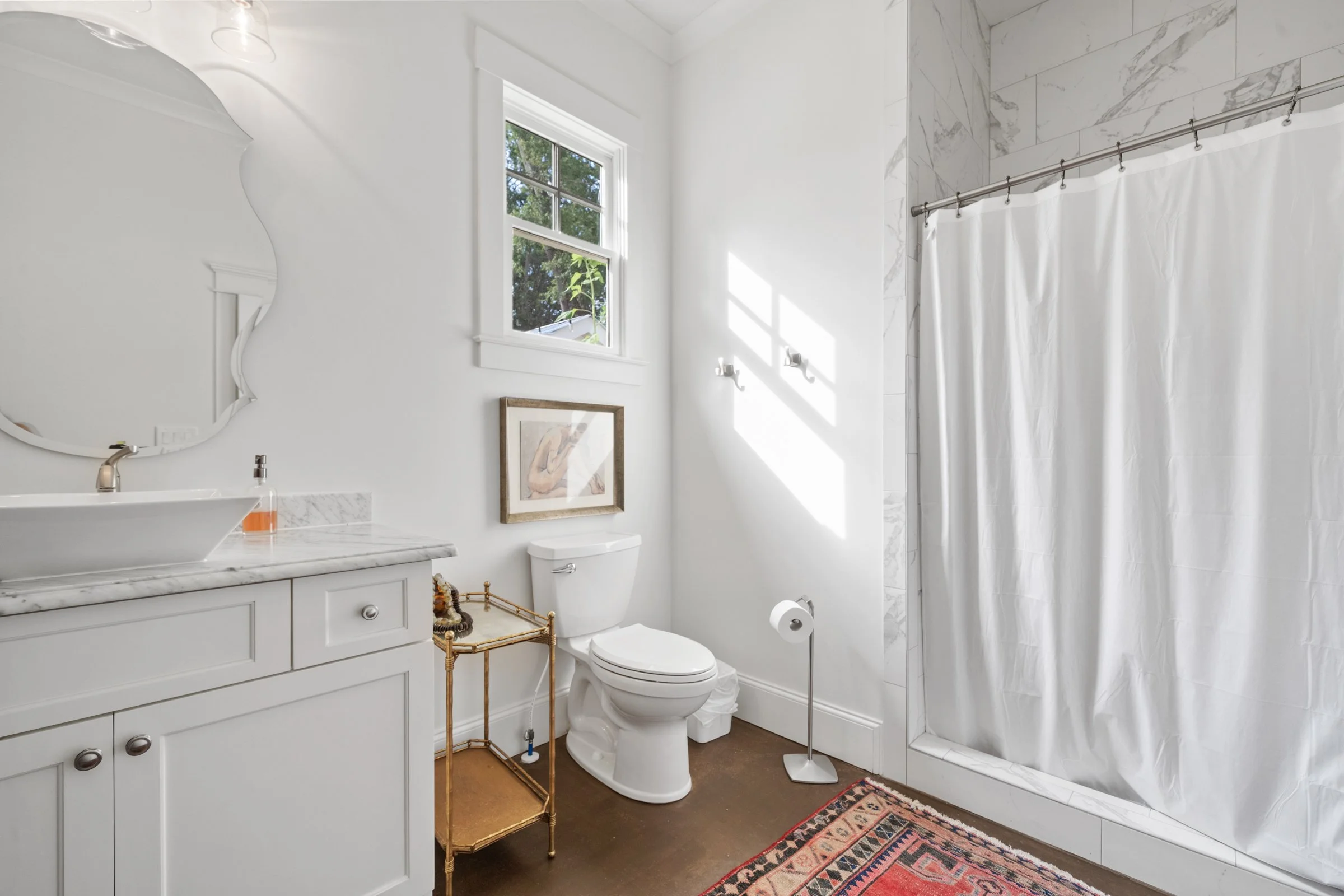 Bright bathroom featuring a white toilet, a marble countertop with a vessel sink, a decorative gold side table, a framed artwork, a window with natural light, a white shower curtain, and a patterned rug.