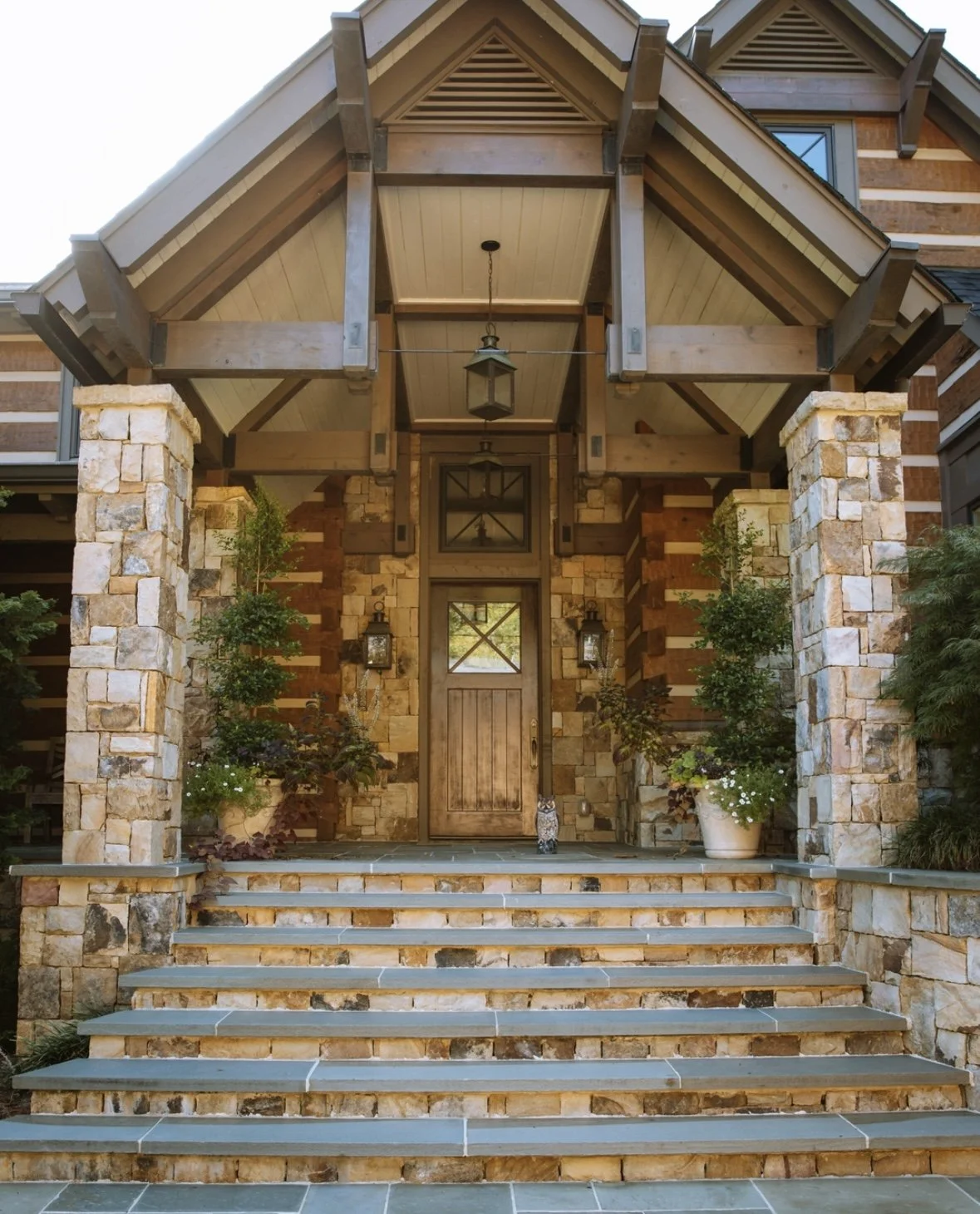 Front entrance of a house with stone pillars, wooden door, stairs, and potted plants.