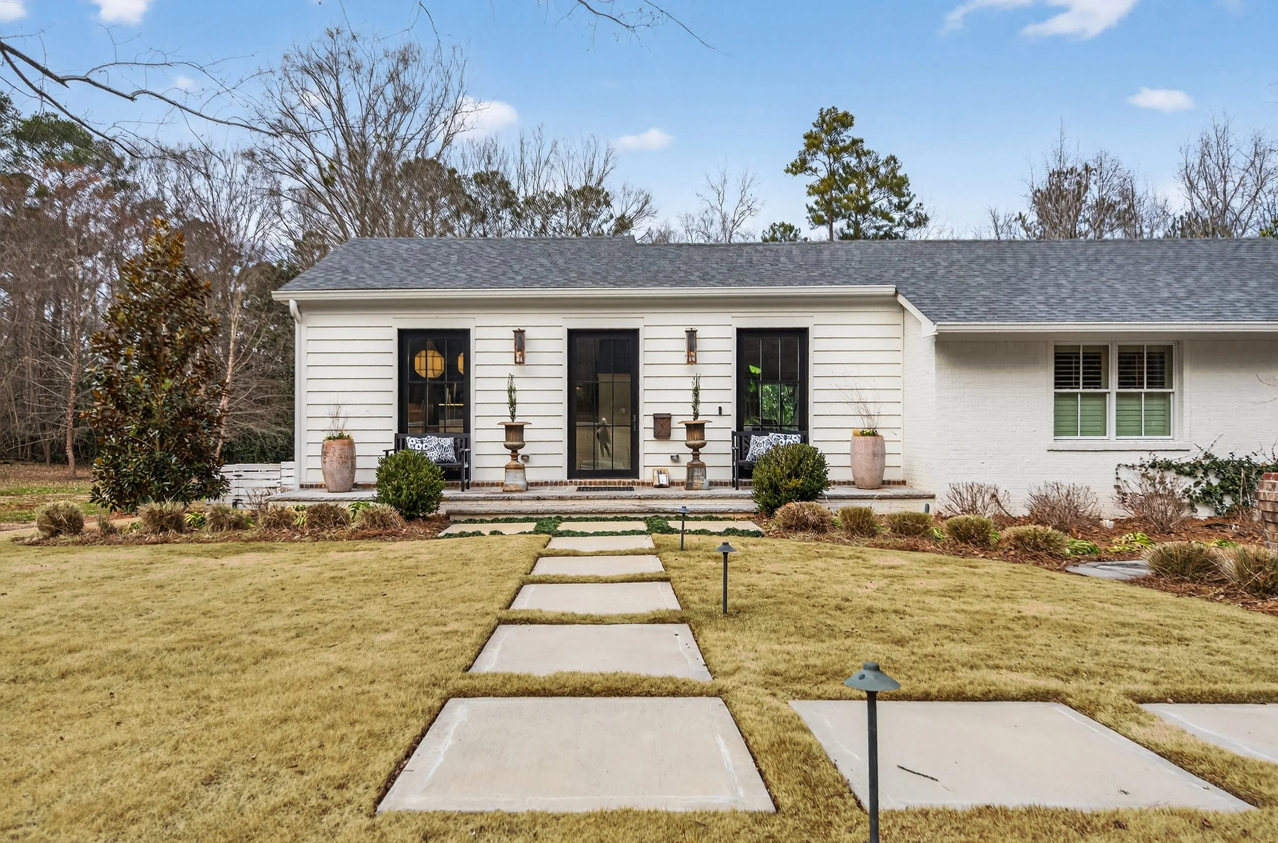 Front view of a white house with a gray shingled roof, black window frames, and a small porch with concrete steps. The porch has potted plants, a bench, and decorations. The yard features a stone pathway, grass, shrubs, and lawn lights, with trees in