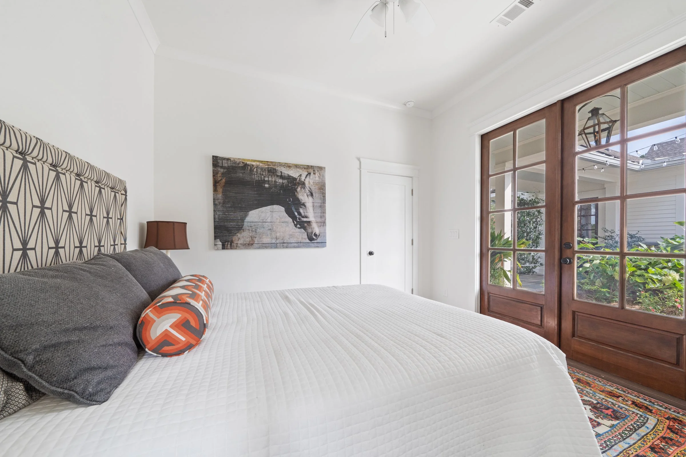 Bright bedroom with large bed, brown headboard, gray pillows, orange patterned bolster pillow, white quilt, wooden sliding door with glass panels, white walls, ceiling fan, artwork of horse, colorful rug, and views of garden outside.