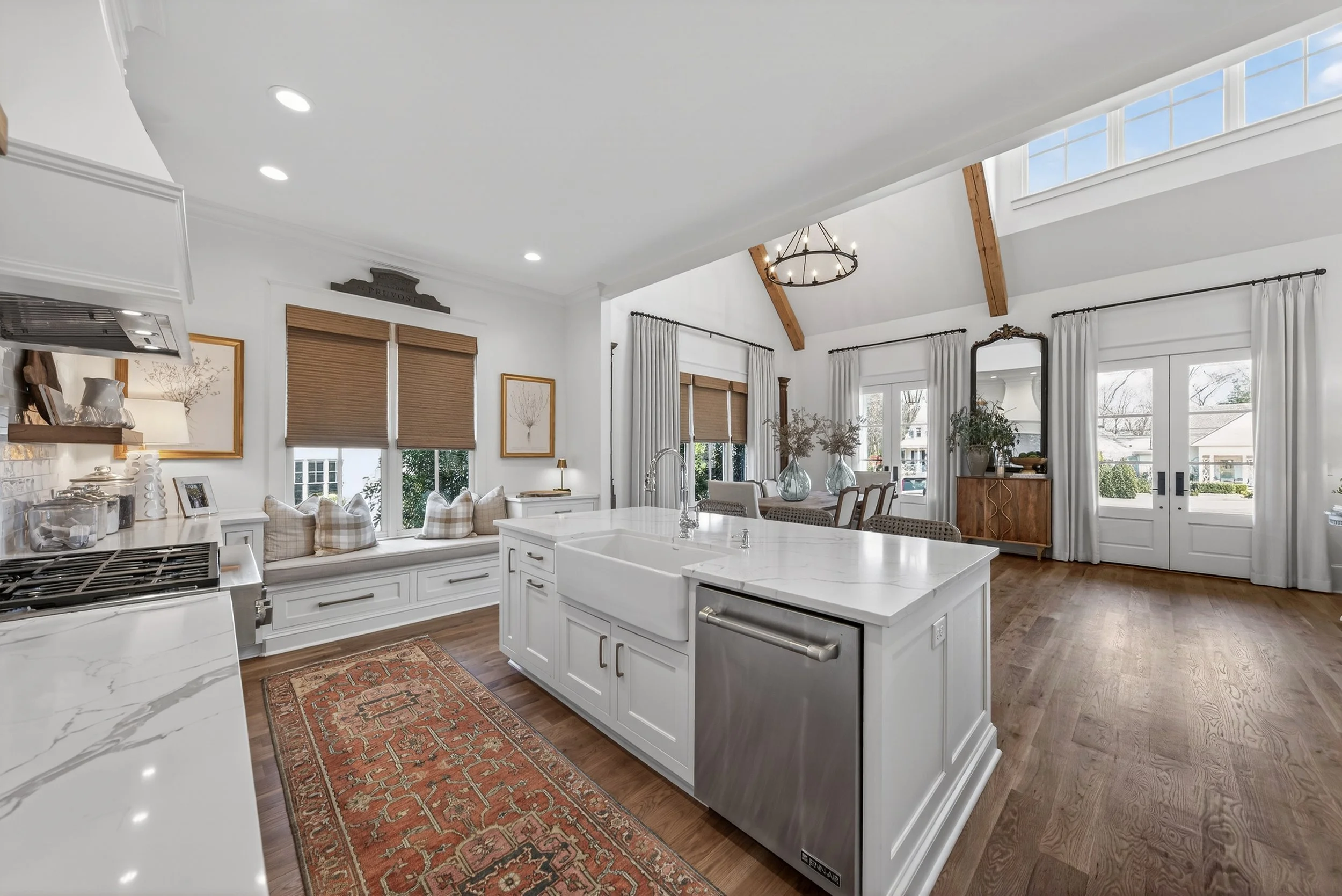 Bright, open kitchen and living space with white cabinetry, a large kitchen island with a farmhouse sink, wooden flooring, and large windows with curtains and blinds, decorated with plants and vases.