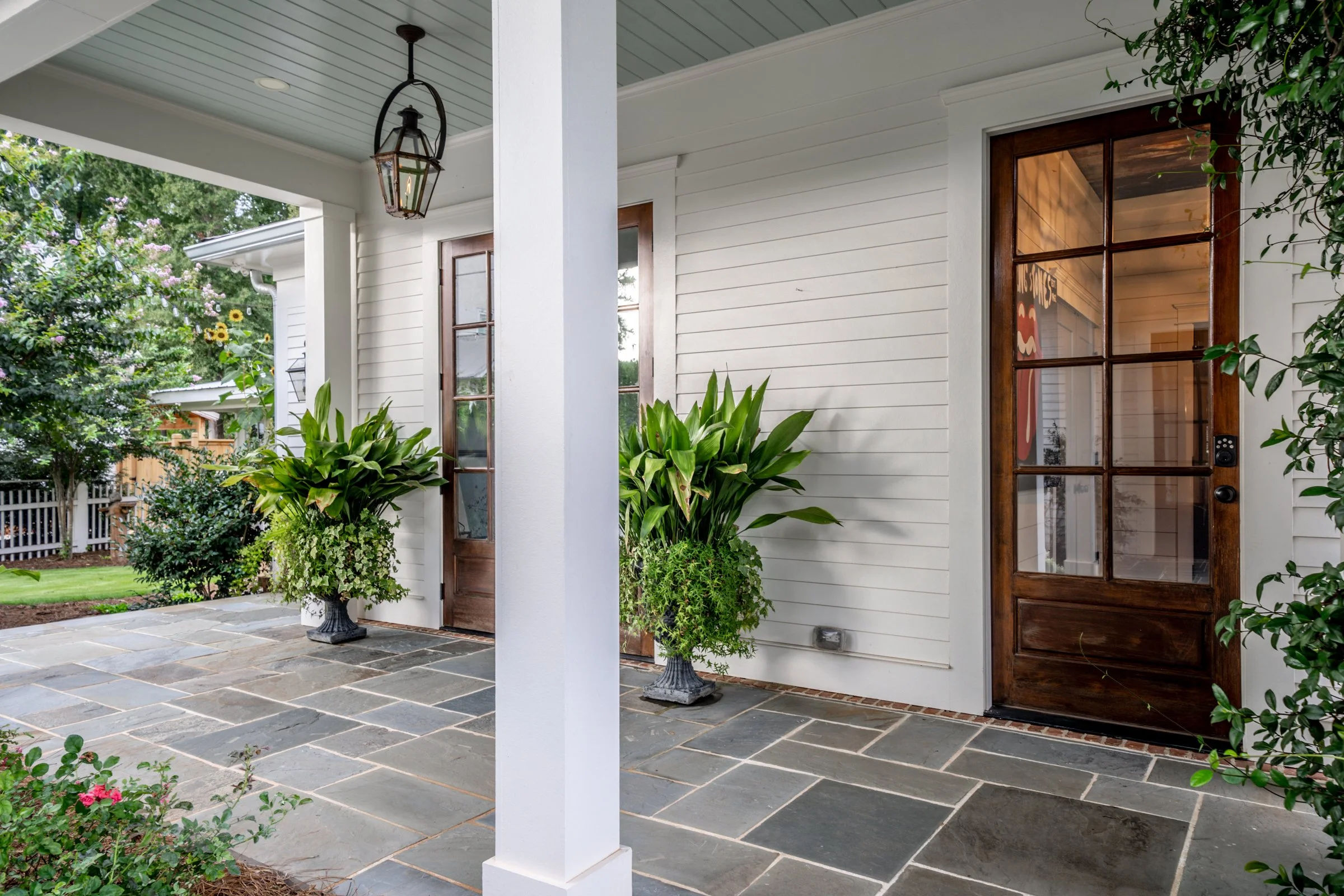 Front porch of a house with white siding, large potted plants, and a wooden door with glass panels, surrounded by green plants and trees.