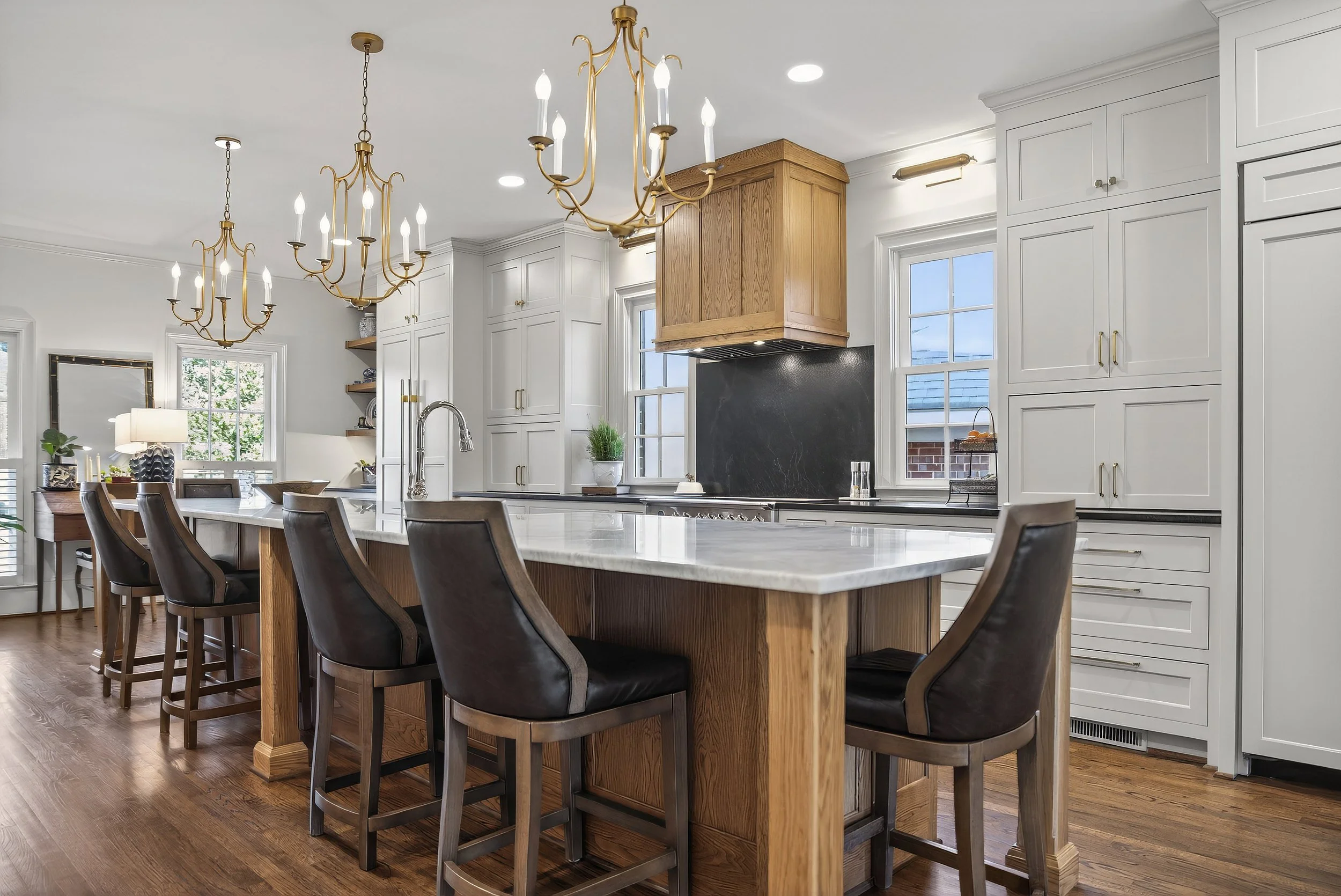 Modern kitchen with white cabinetry, a large island with a marble countertop, wooden accents, black leather chairs, and brass chandeliers.