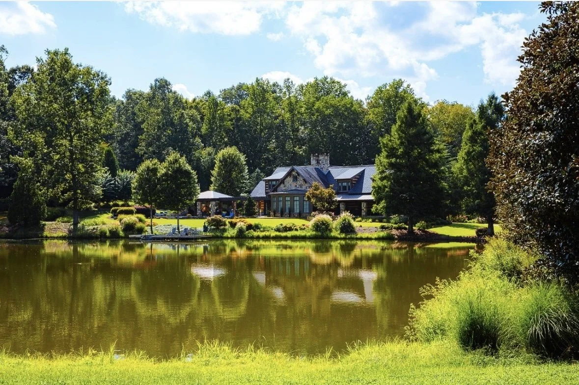 A house on a lush green lawn beside a calm pond, surrounded by trees and a clear blue sky.