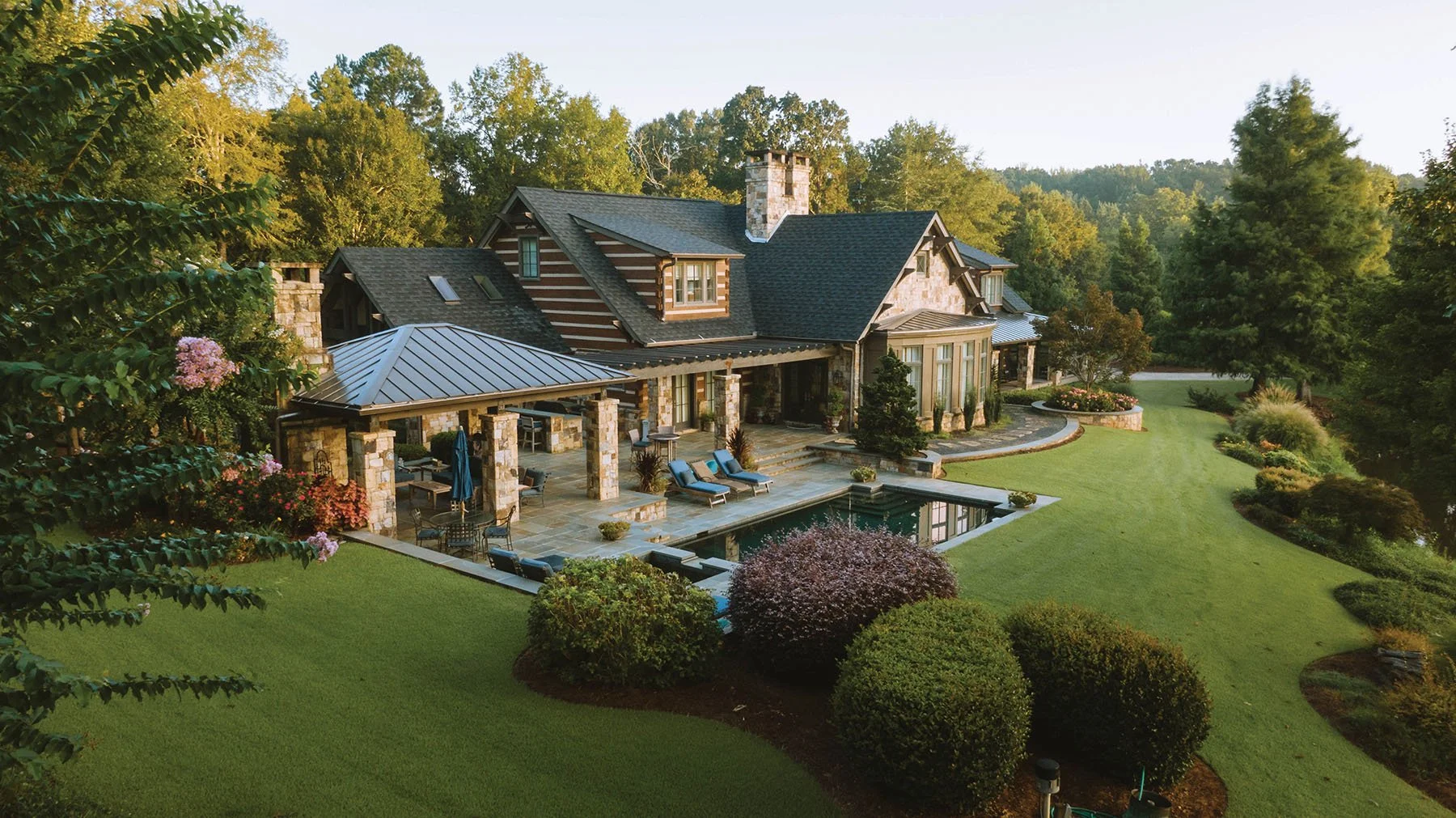 A large home with a stone and wood exterior, multiple rooflines, and a chimney. The backyard features a patio with seating, a small pool, well-maintained lawn, and landscaped bushes and trees, surrounded by tall trees and a hilly landscape.