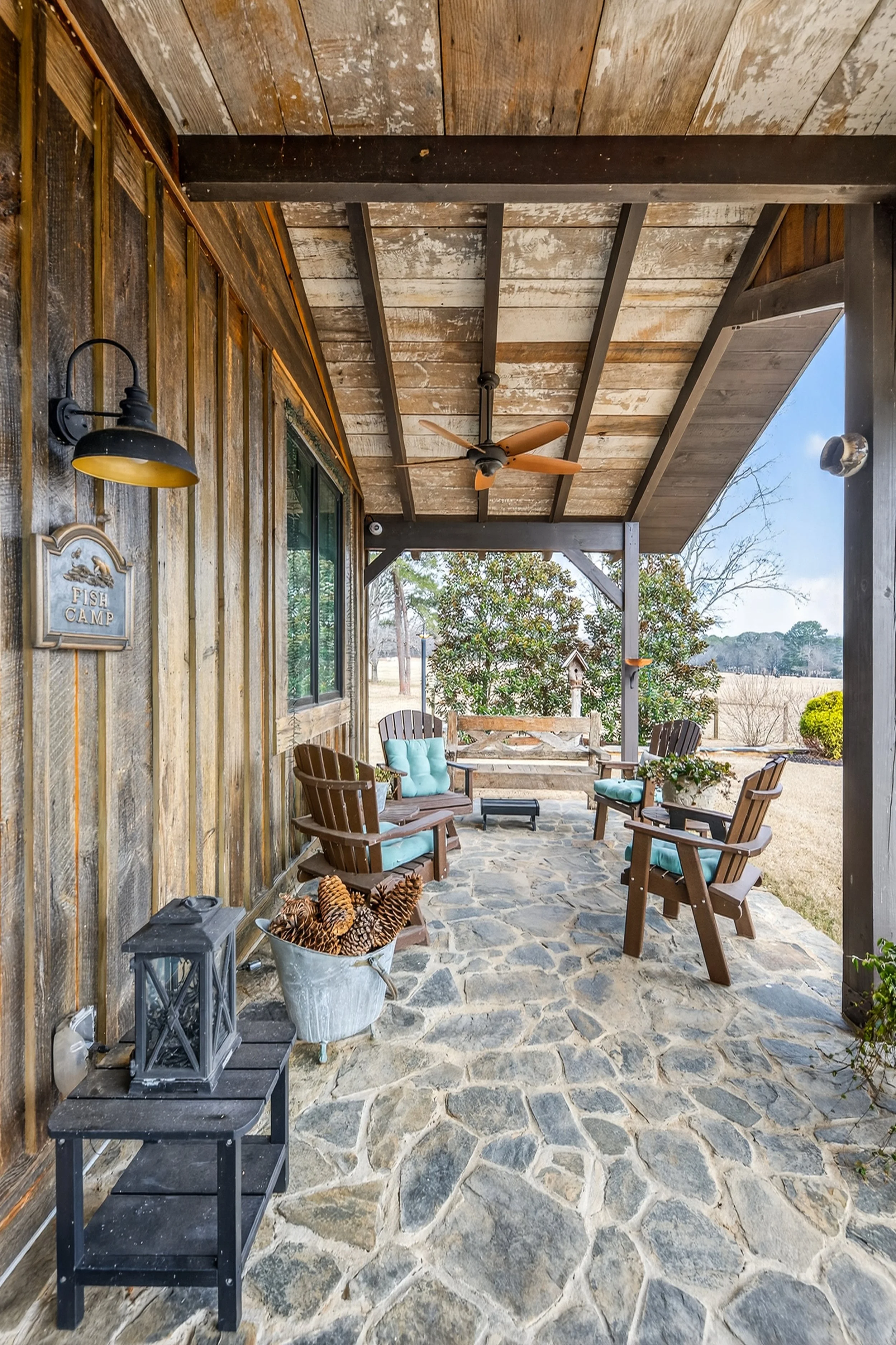 Covered porch with patio furniture, including Adirondack chairs with blue cushions, a bucket of pinecones, and a lantern, on a stone floor, with trees and a field in the background.