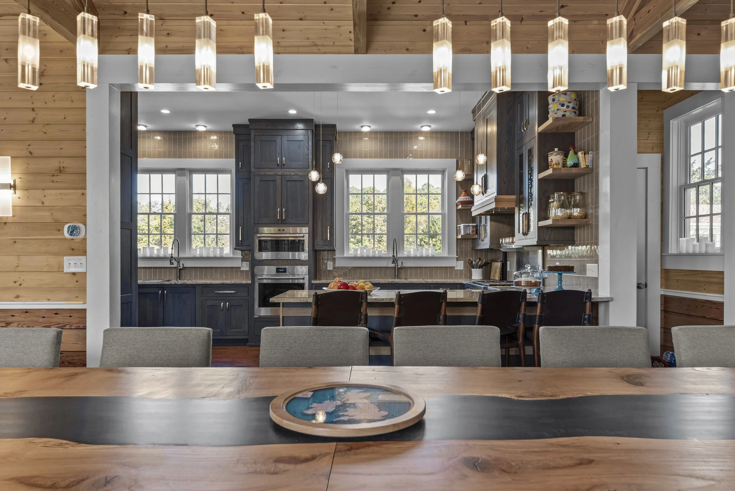 Kitchen with dark cabinets, large windows, island with chairs, modern lighting, and a dining table in the foreground.