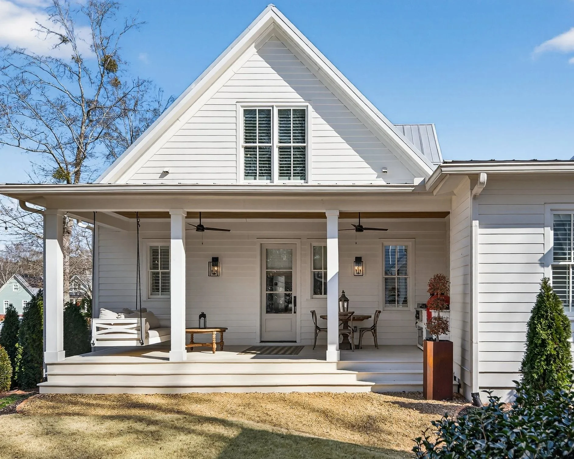 White house with a front porch, ceiling fans, outdoor seating, potted plants, and large windows, under a clear blue sky.