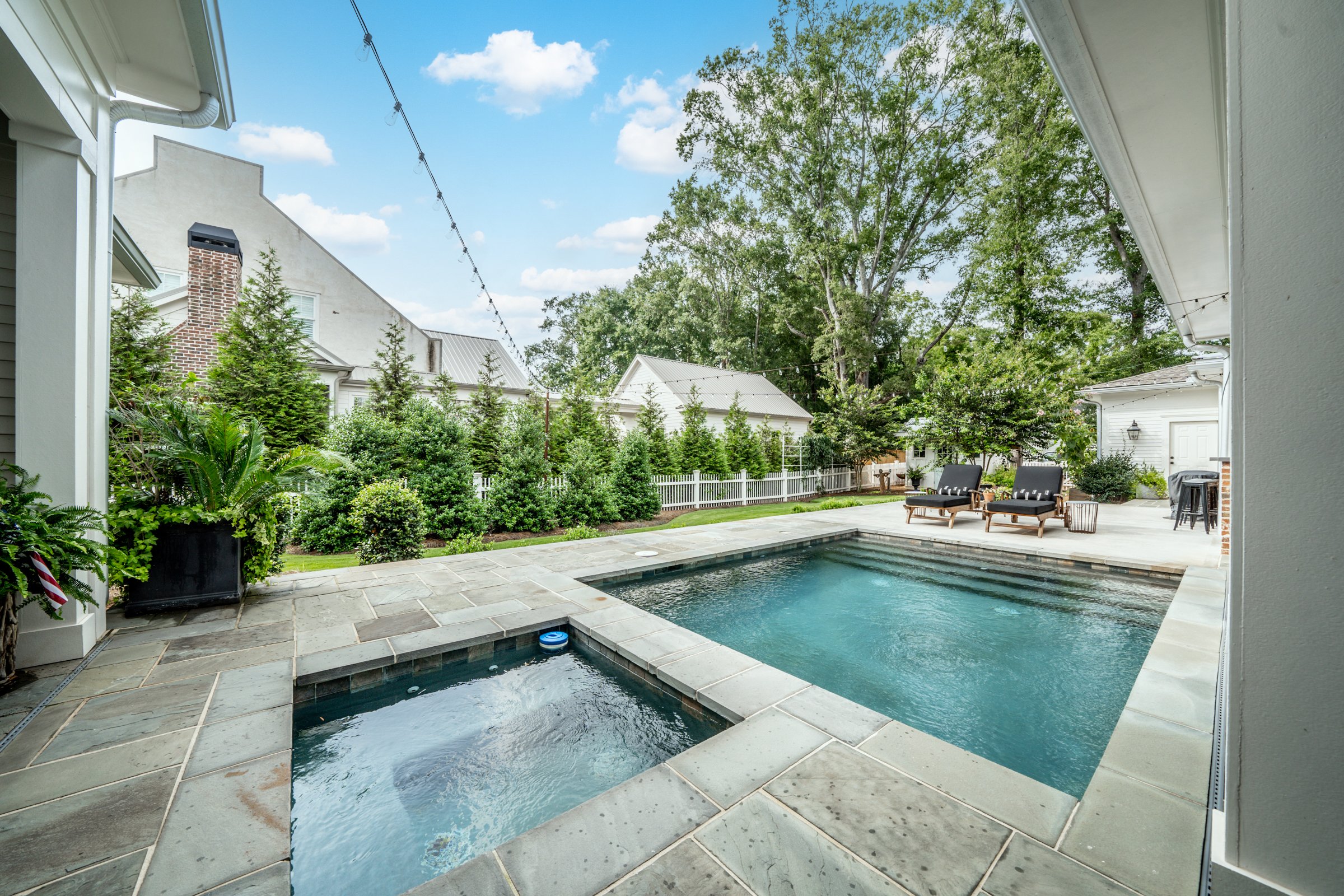 Backyard with a private pool and hot tub, surrounded by greenery and outdoor furniture, under a partly cloudy sky.