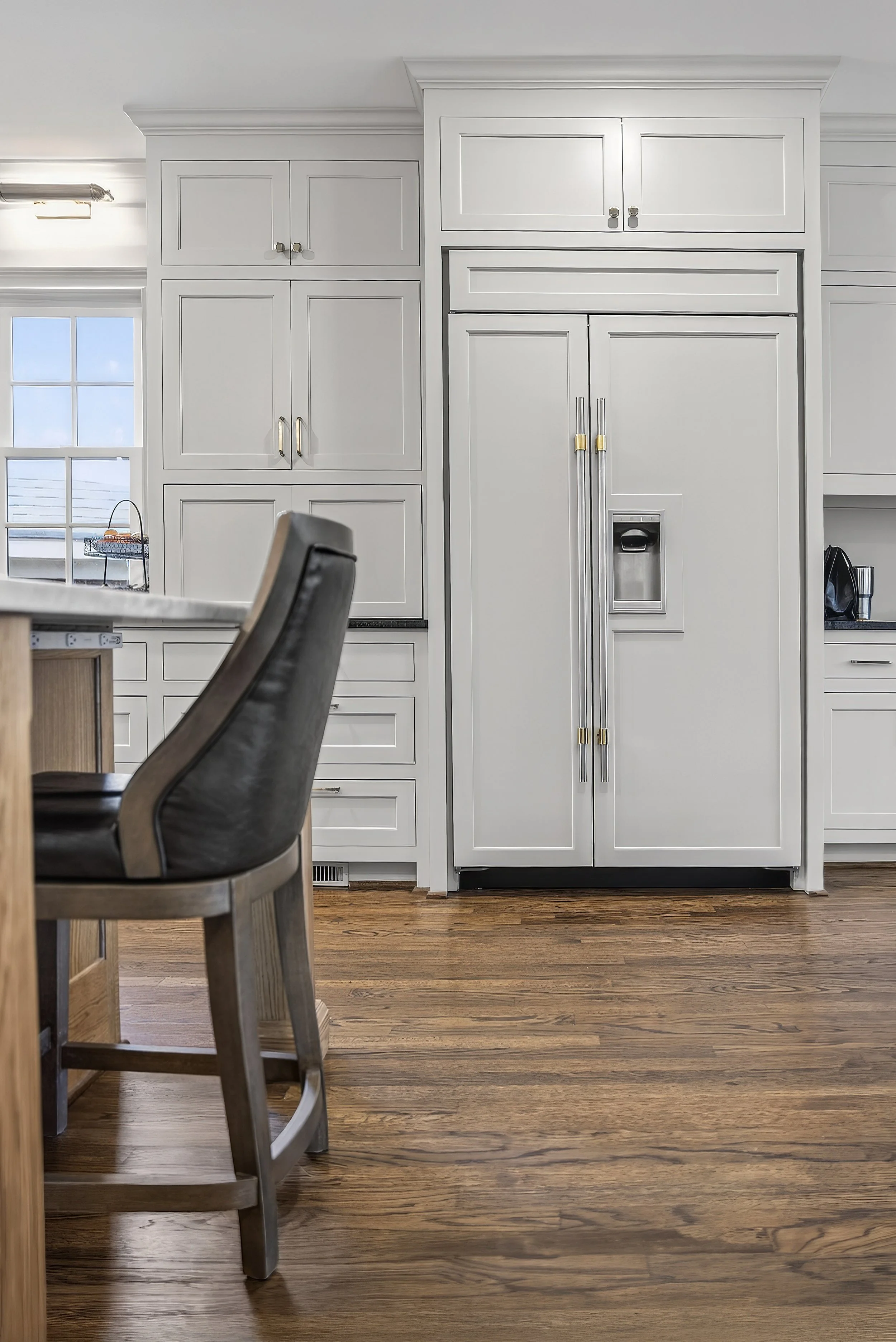 White kitchen with a large stainless steel refrigerator, white cabinets, and hardwood floors. A window is visible on the left side.