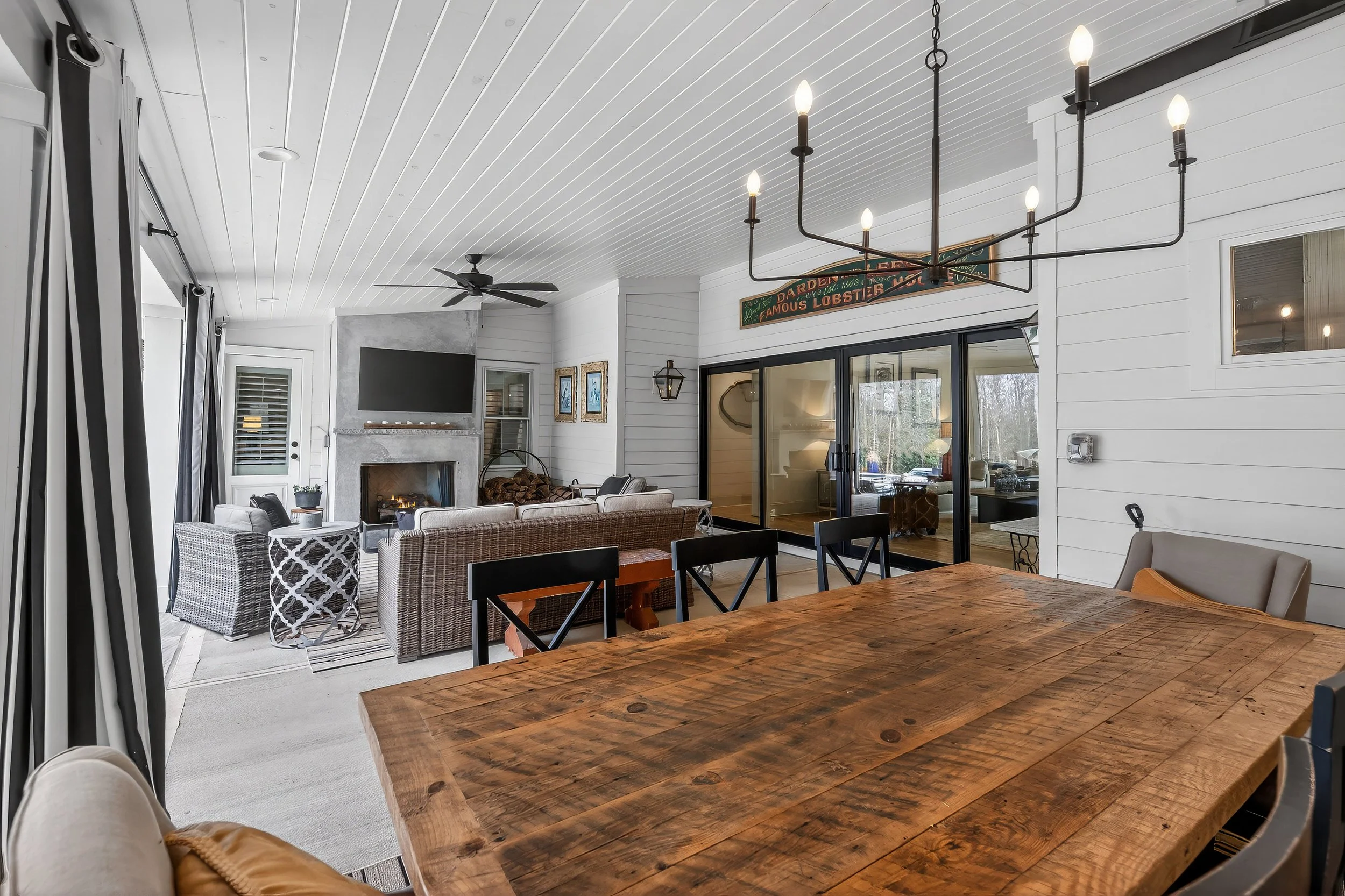 Interior view of a dining and living area with a wooden dining table, black chairs, a fireplace, a large flat-screen TV, a sofa, and decorative wall art, with light-colored wood paneling and sliding glass doors.