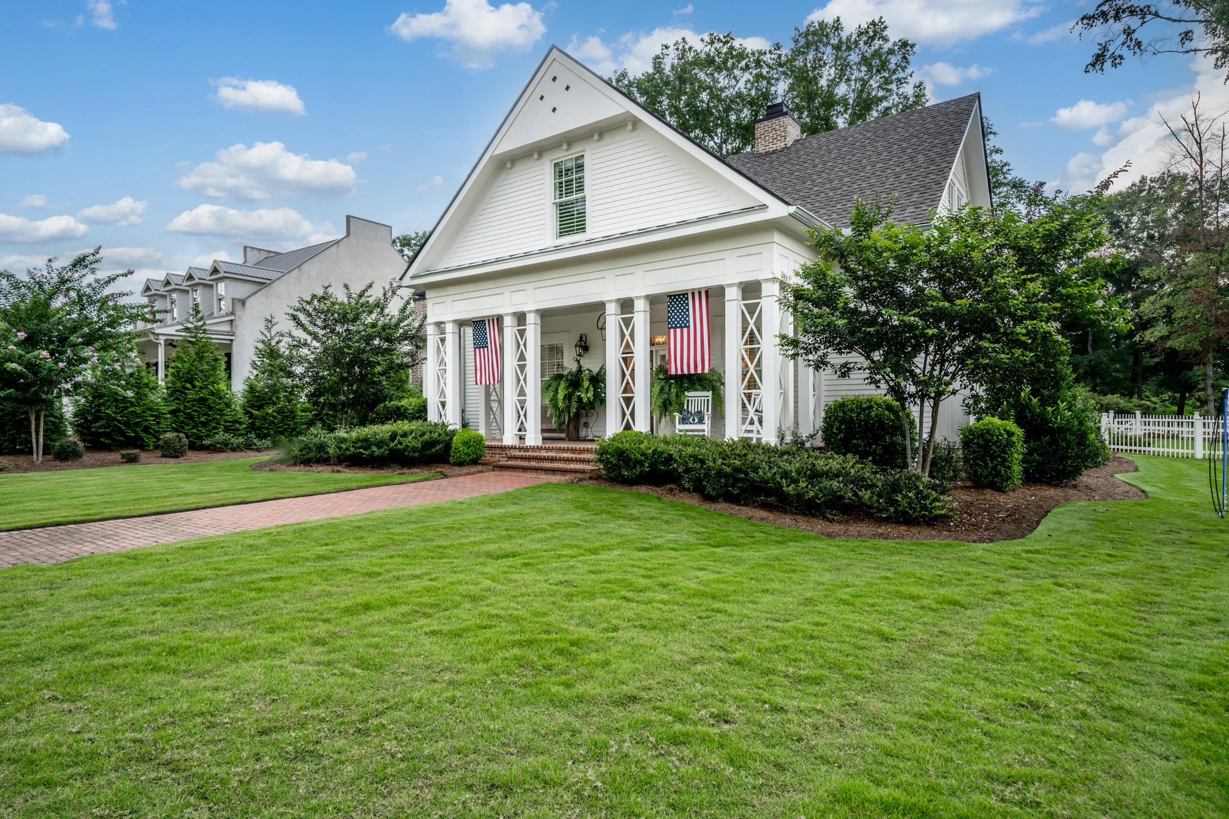 A white house with a large front porch, decorated with American flags and green plants, surrounded by a neatly manicured lawn and trees under a partly cloudy sky.