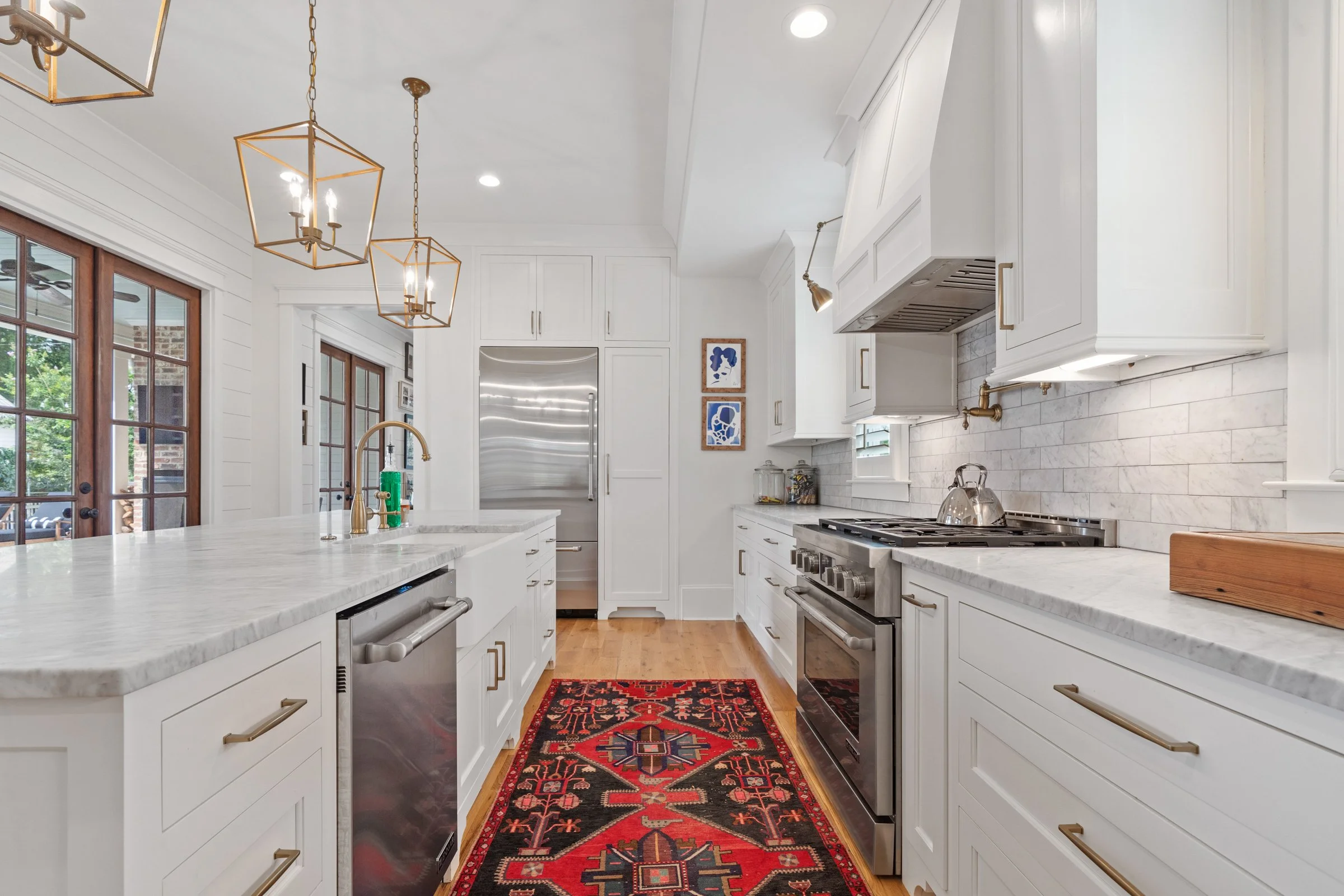 Bright, modern kitchen with white cabinets, marble countertops, stainless steel appliances, wooden flooring, and a red patterned rug. Gold hardware and lighting fixtures add elegance.