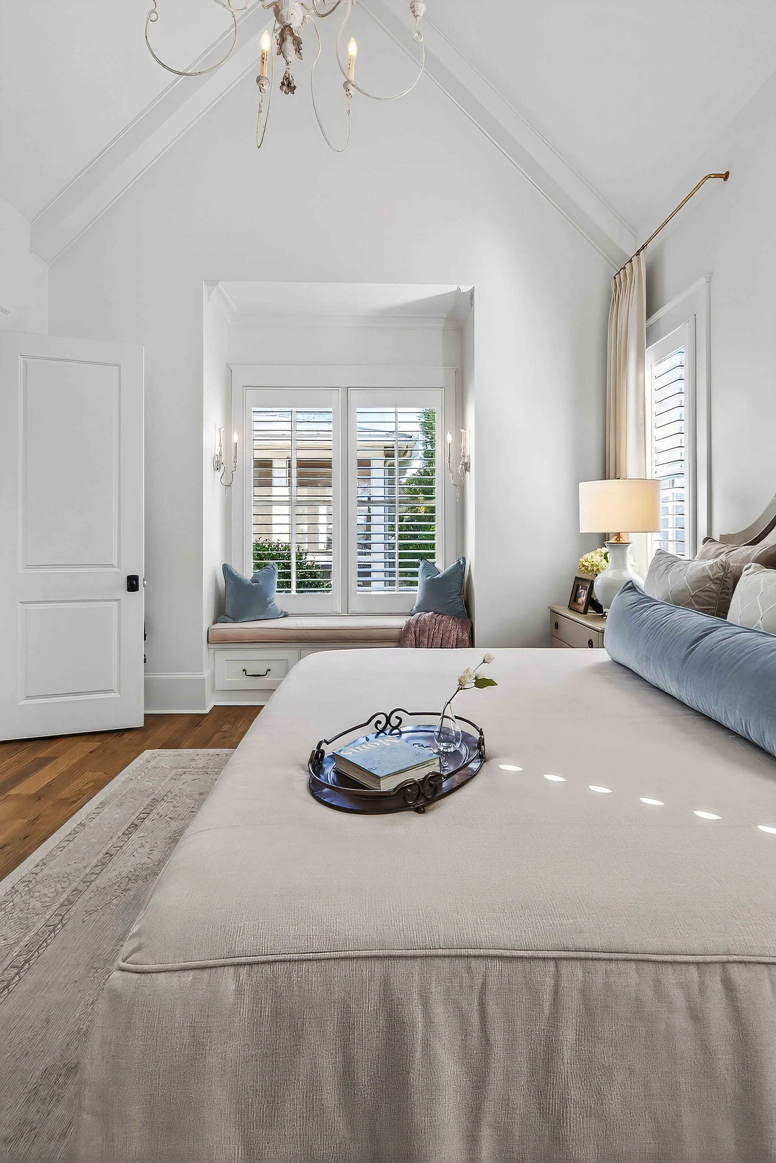 Bright bedroom with white walls, large bed with light-colored bedding, tray with book and flower on the bed, window seat with pillows, window with shutters, chandelier, and side table with lamp.