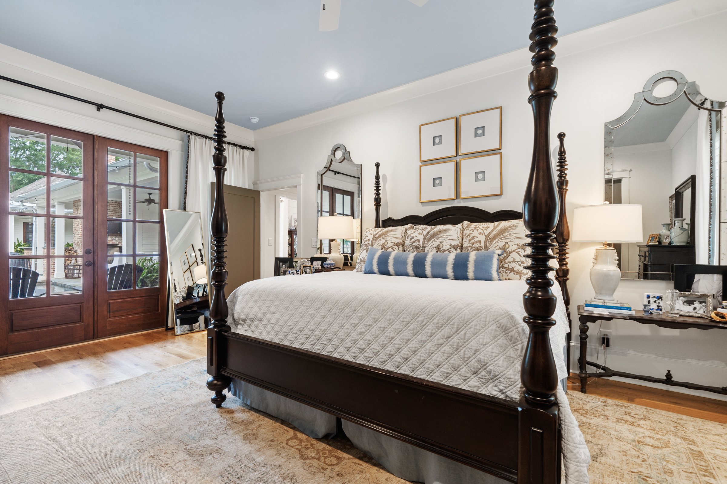 A bedroom with a dark wooden four-poster bed, white bedding, and patterned pillows. There are two nightstands with lamps, a mirror, and framed wall art. French doors lead outside, and a small dresser is visible in the corner.