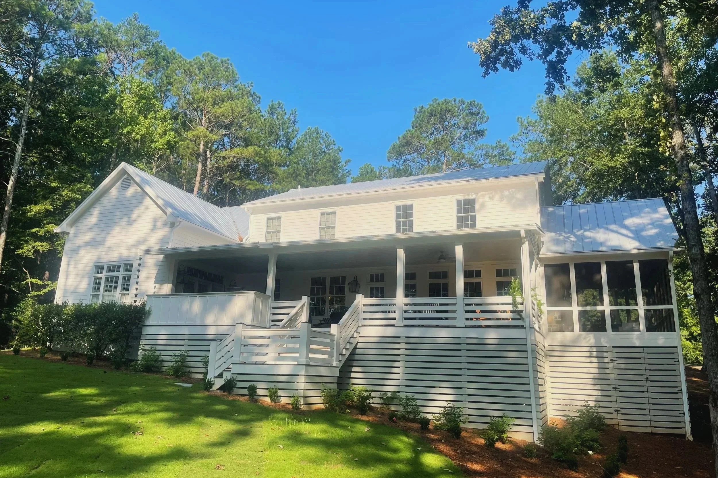 A white two-story house with a screened porch, set on a grassy lawn surrounded by trees with blue sky overhead.