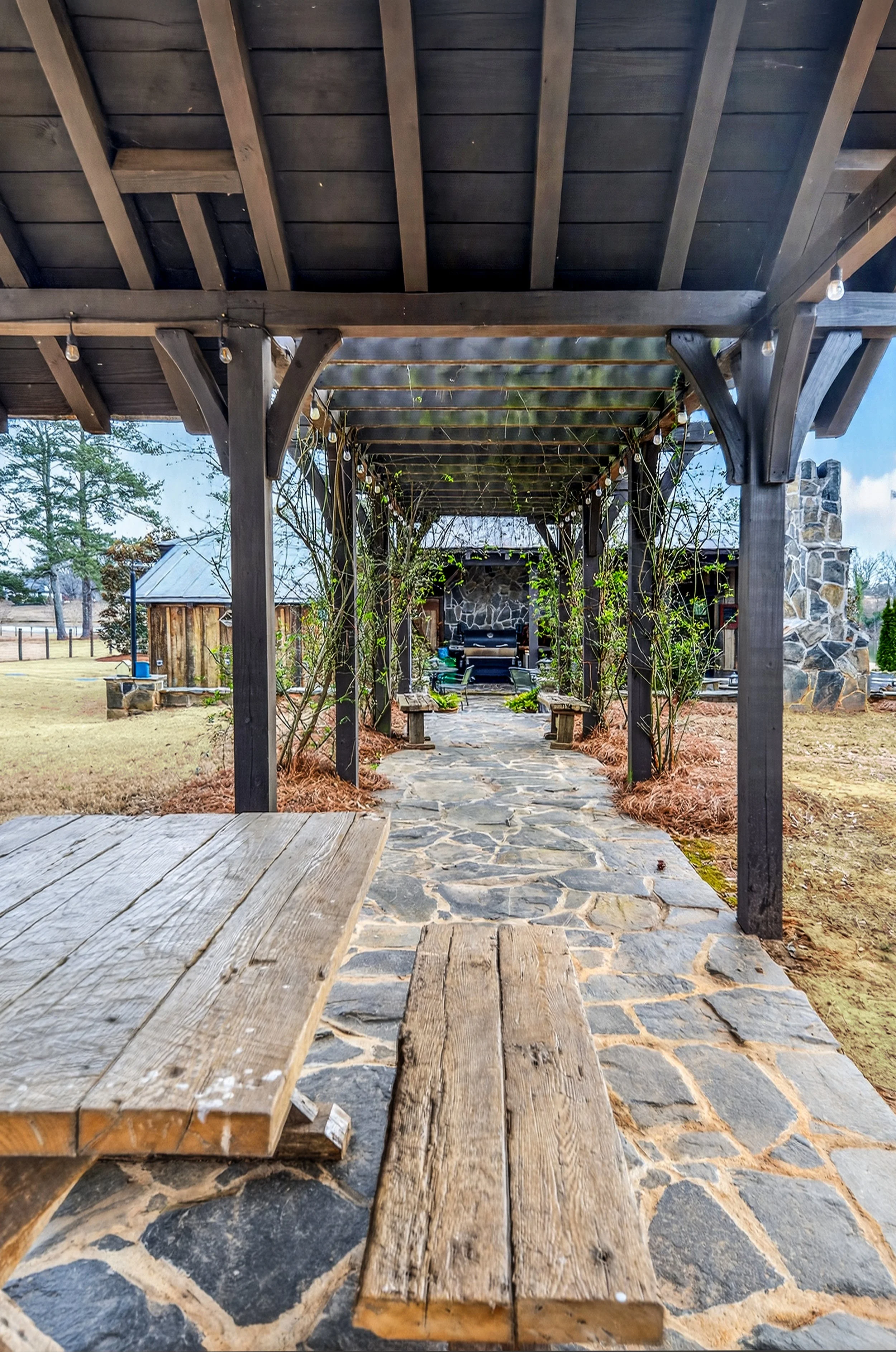 A stone pathway under a wooden pergola with string lights, leading to an outdoor seating area with a stone fireplace and benches.