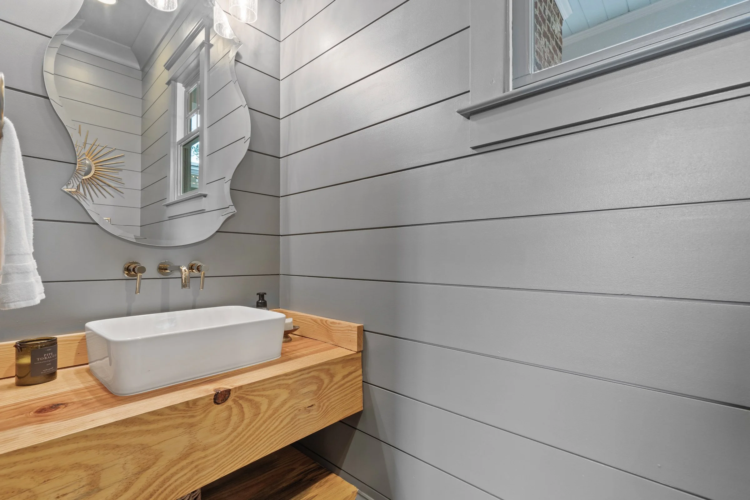 Modern bathroom with gray shiplap walls, a wooden vanity with a rectangular vessel sink, a decorative round mirror, and a window with a white frame.