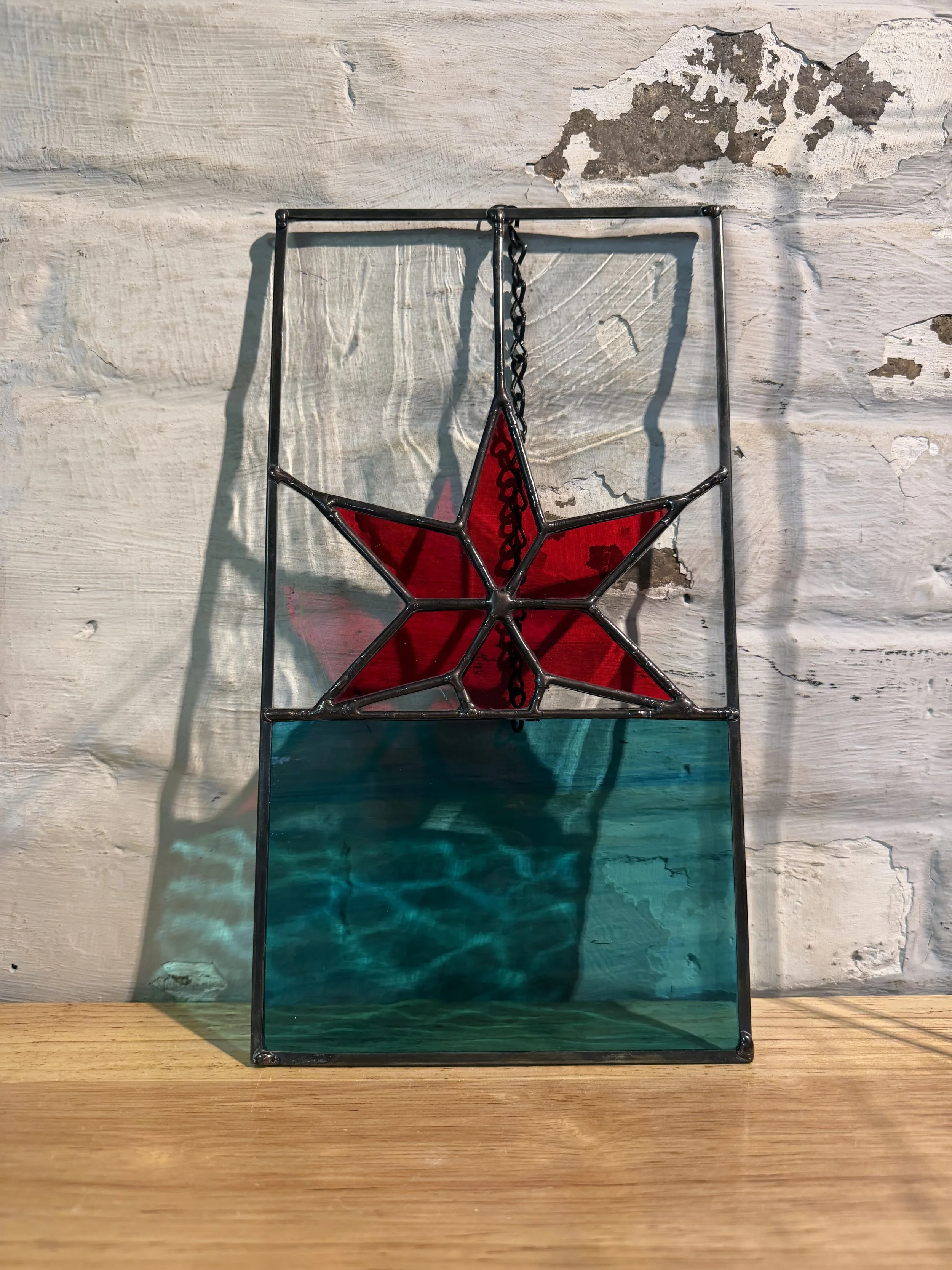 Decorative glass panel with a red star and teal background, mounted on a black metal frame, leaning against a worn white wall.