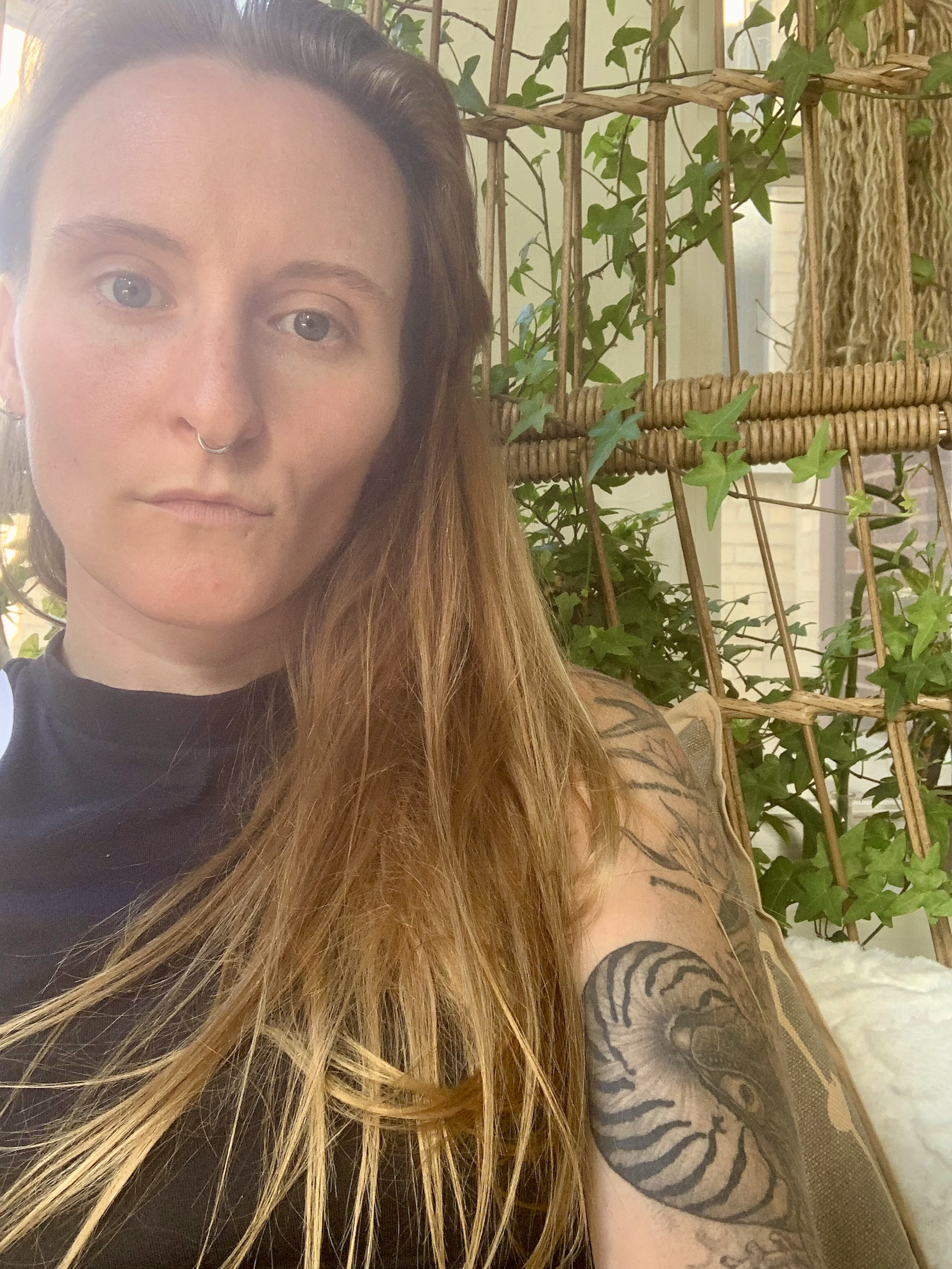 A woman with light skin, long red hair, and a septum piercing, sitting in front of a wooden lattice with green ivy plants, looking at the camera.