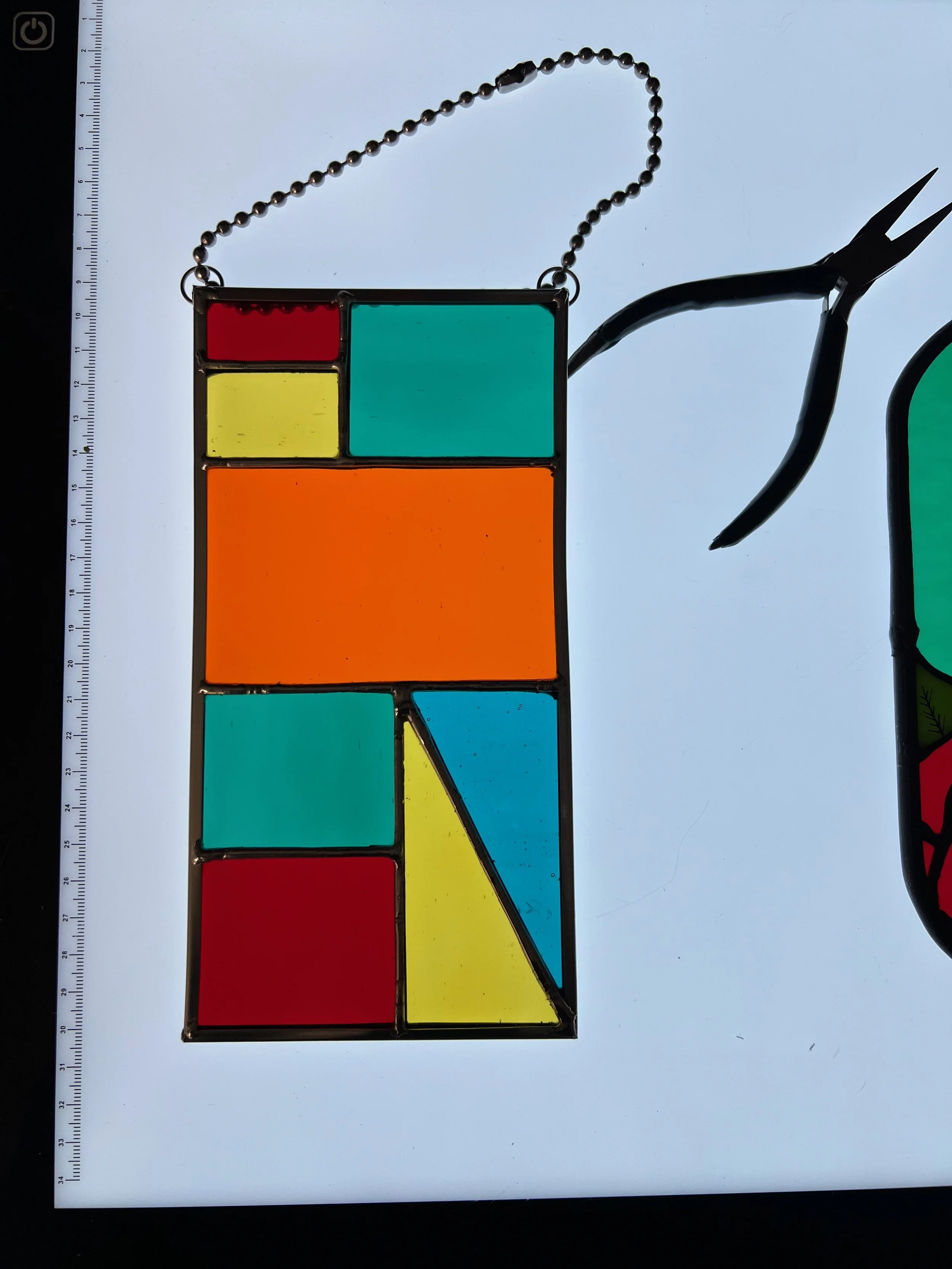 Colorful stained glass stained glass panel with geometric shapes and a small, red, yellow, teal and orange rectangle and a blue triangle, plus a black pair of pliers with a curved handle and pointed tip.