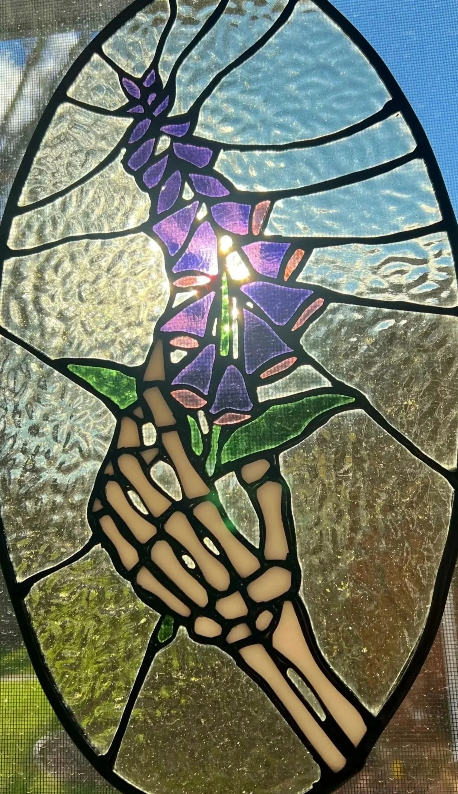 A stained glass window depicting a human hand holding a purple flower with green leaves against a textured glass background.