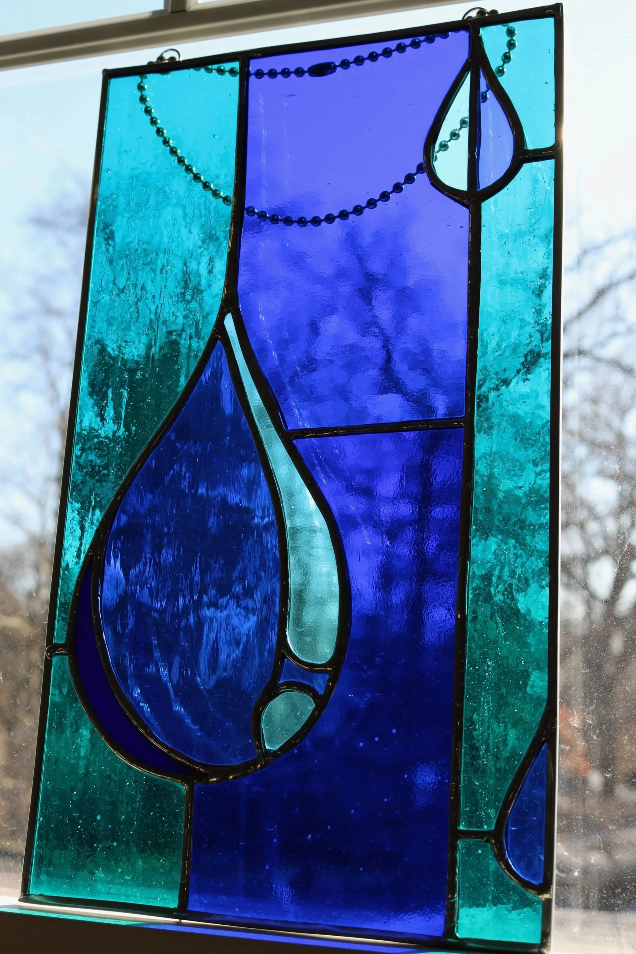Colorful stained glass window featuring various blue and teal shapes with black outlines, hung in front of a window with a background of a cloudy sky and leafless trees.