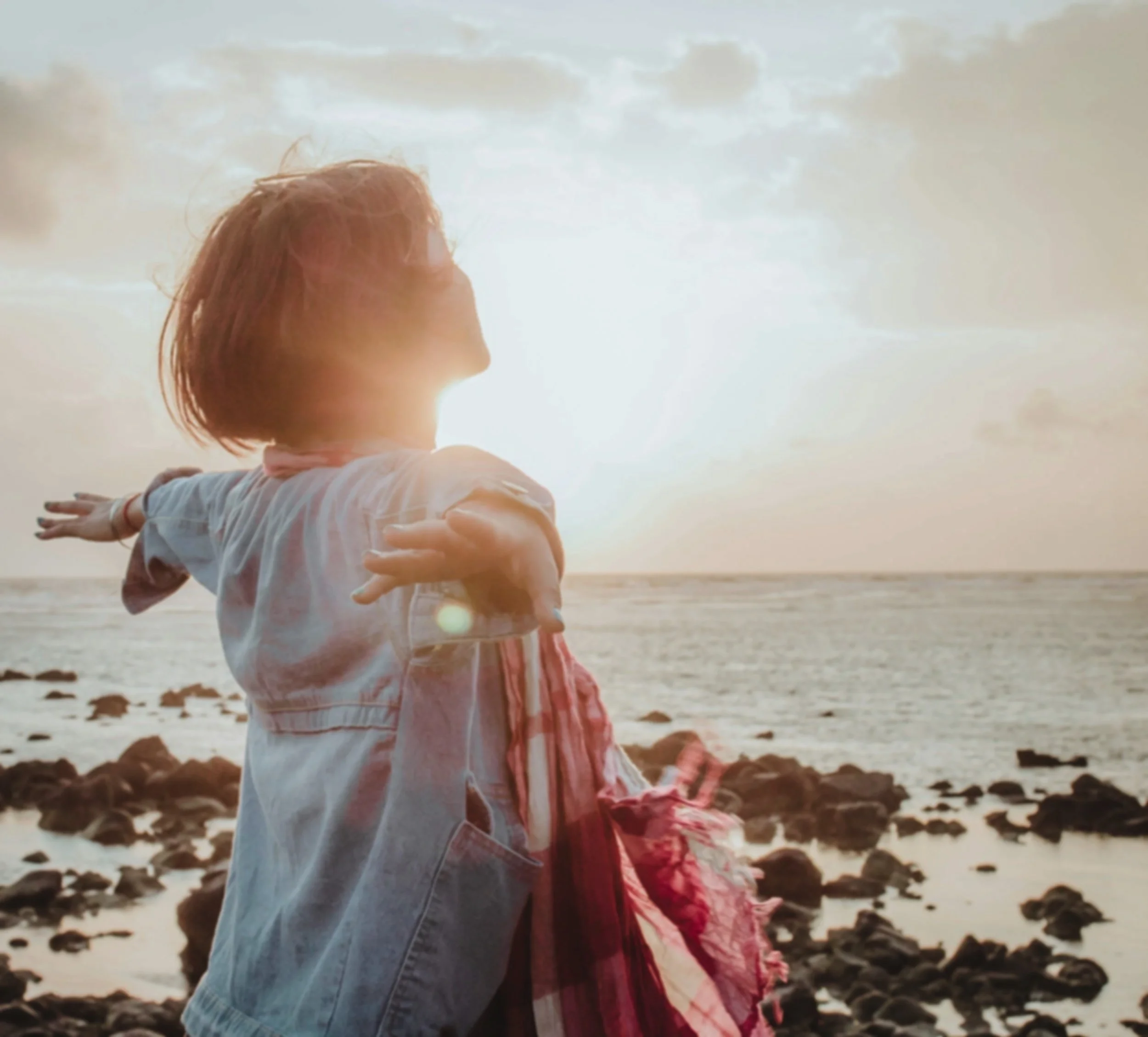 A woman and a child standing on a rocky beach during sunset, with the woman holding the child's shoulders as they face the ocean.