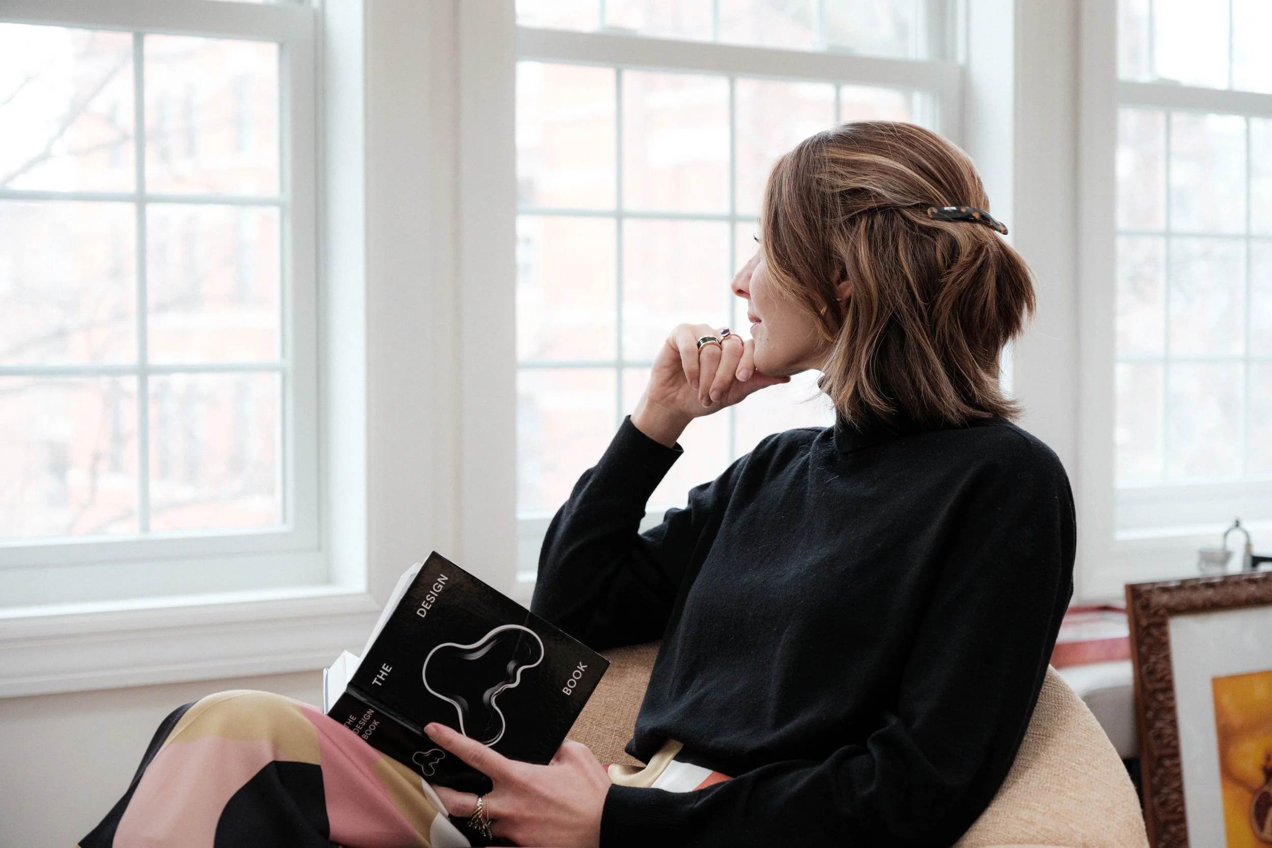 woman sitting in living room chair, holding design book, looking out the window