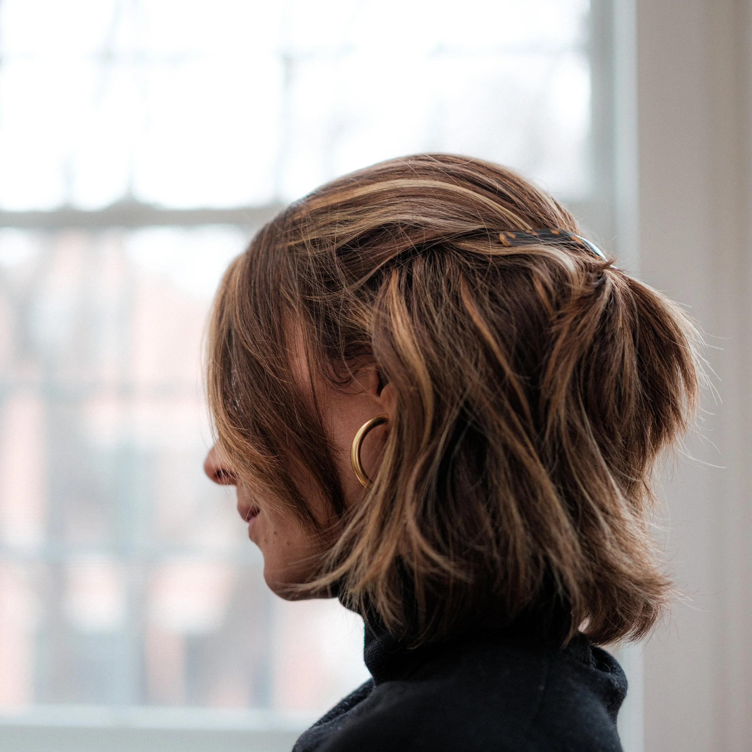 close-up side of woman's face with hair half-up against a living room window backdrop