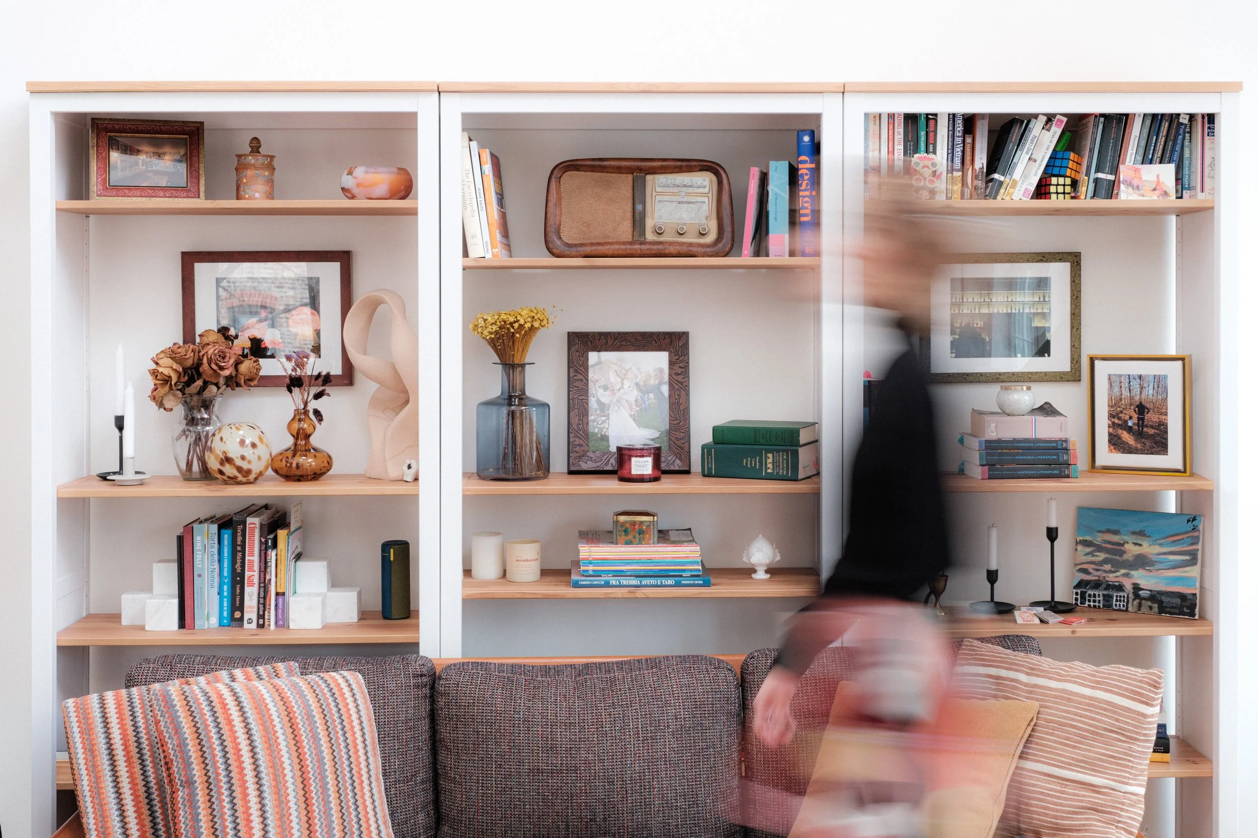 woman walking by living room couch and bookshelves filled with vases, clocks, picture frames, art, and magazines