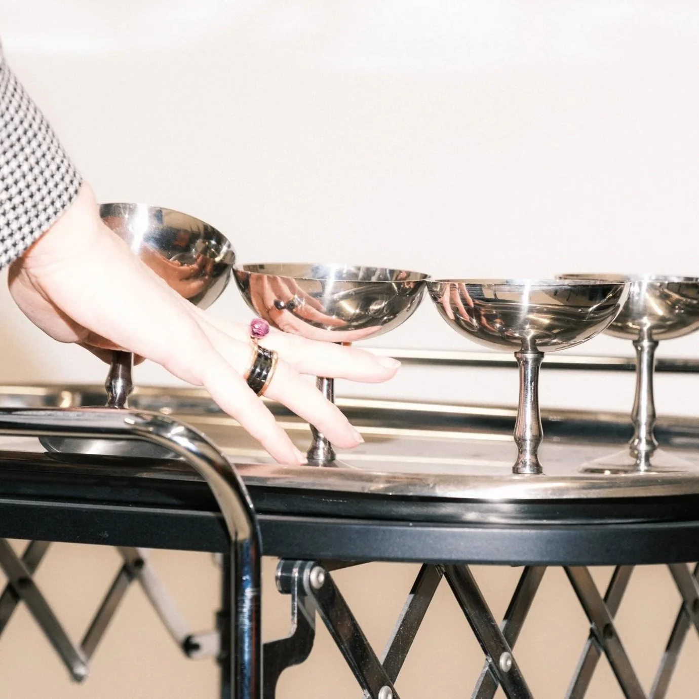 woman's hand picking up silver coupe glasses off of black and silver bar cart against white backdrop