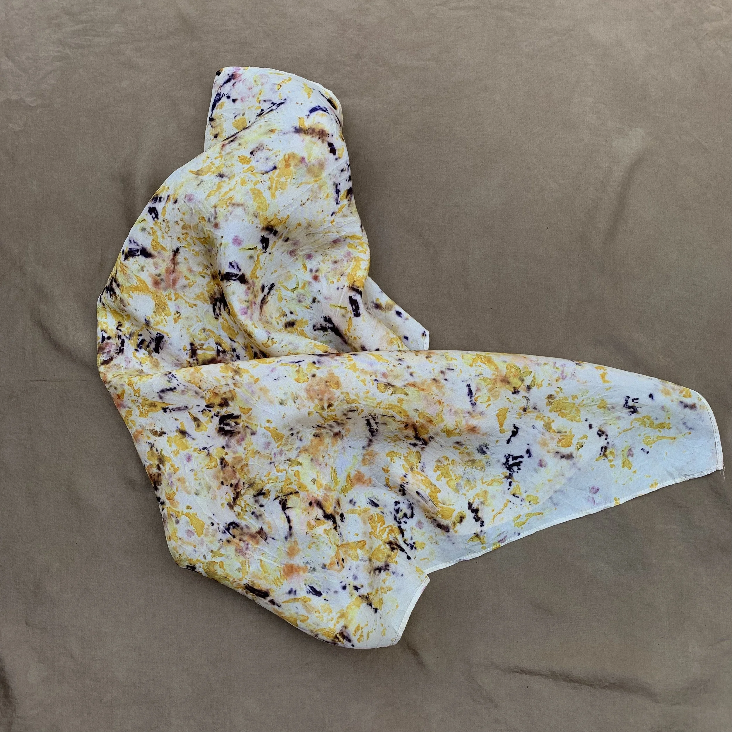 A crumpled white silk scarf with a colorful abstract pattern of yellow, purple, and pink splashes on a plain brown surface.