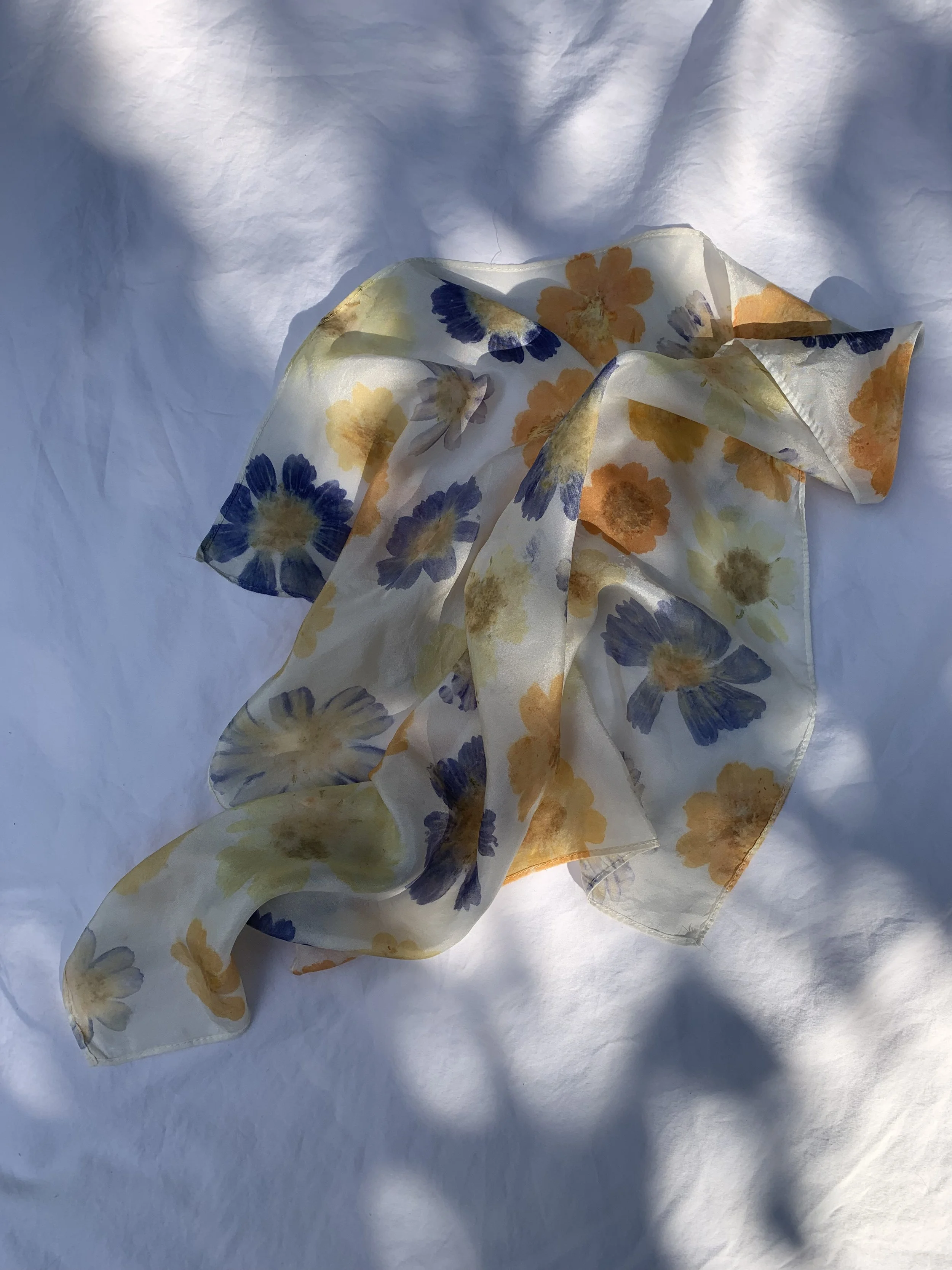 A floral-patterned silk scarf with yellow, orange, and purple flowers on a white background, draped on a white sheet with shadows and light.