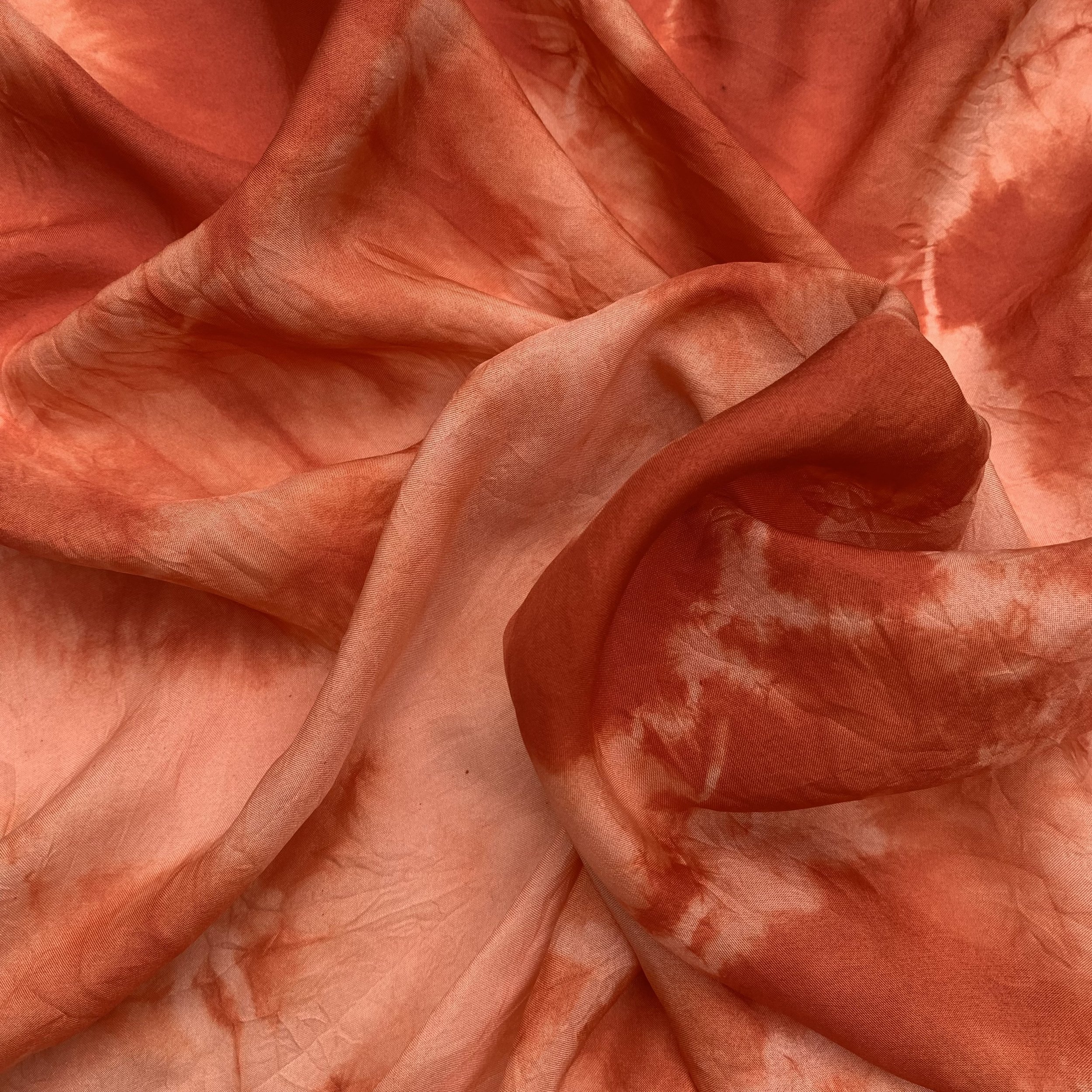 Close-up of a soft, flowing, silk scarf in a redish coral color with some darker and lighter marbled patterns, arranged with gentle folds and ripples.