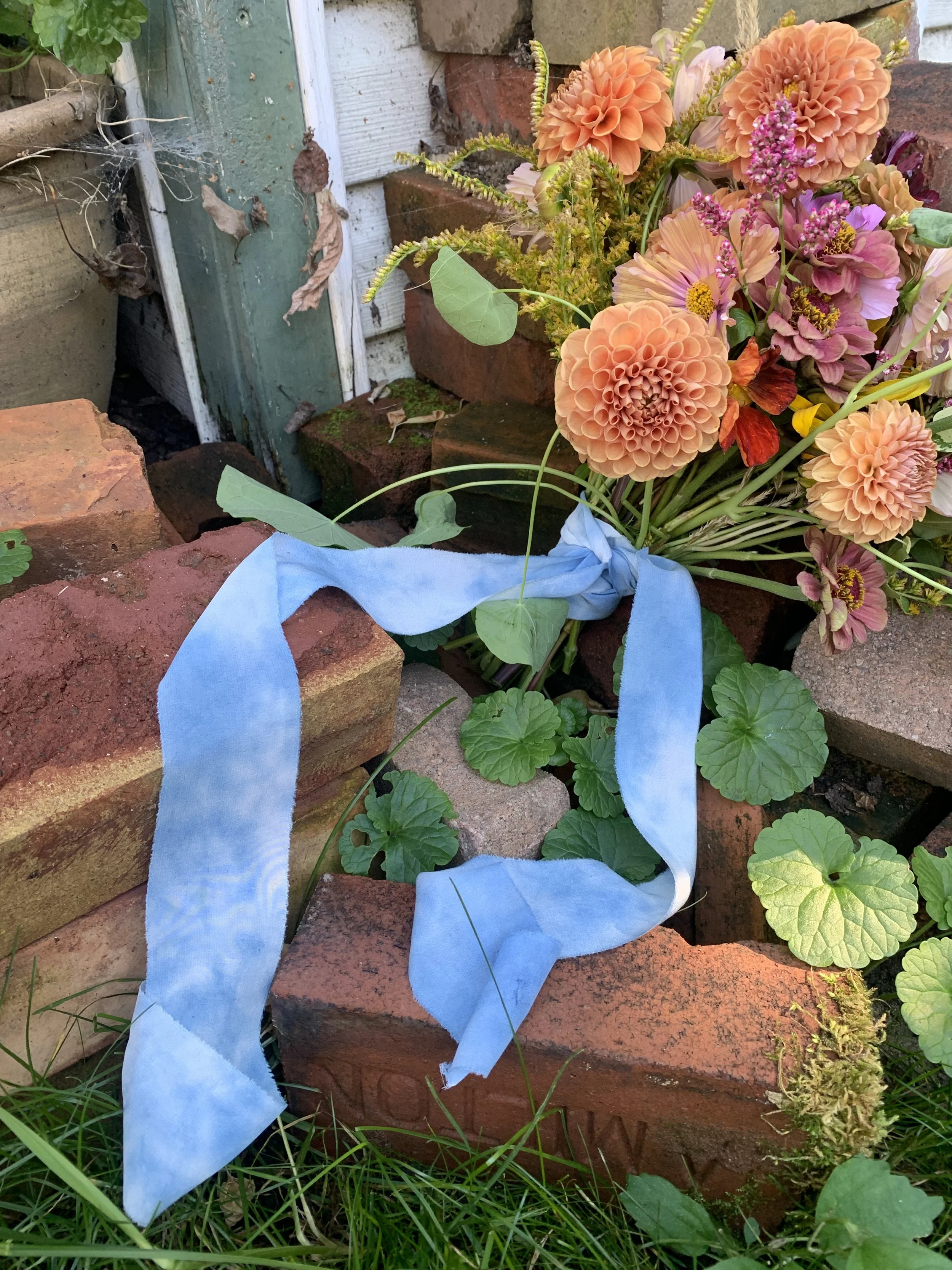 A bouquet of peach and pink flowers tied with a naturally dyed blue ribbon, placed among bricks and green leaves in an outdoor garden setting.