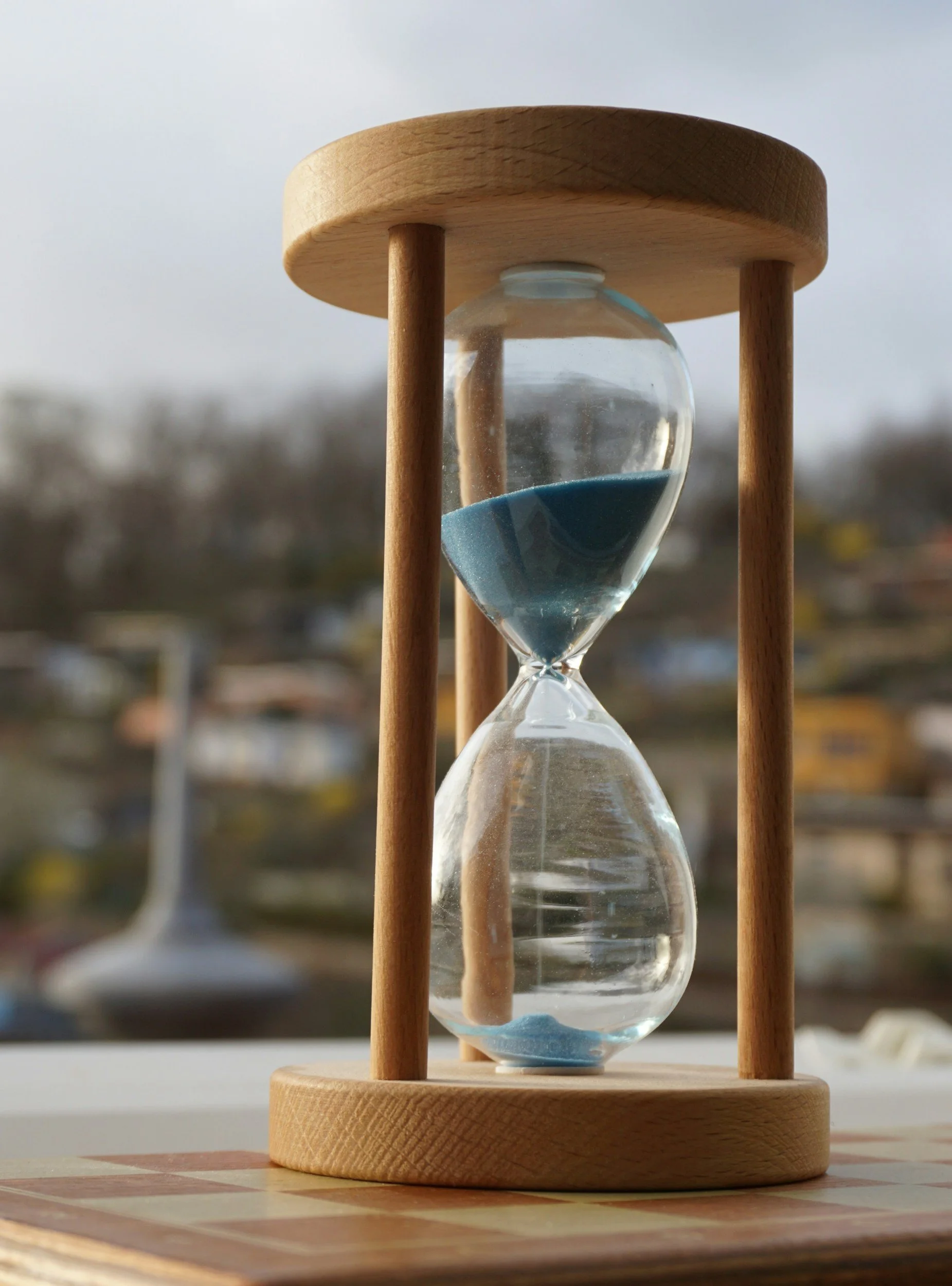 An hourglass with blue sand in a wooden frame on a table, with a blurred background of a cityscape.