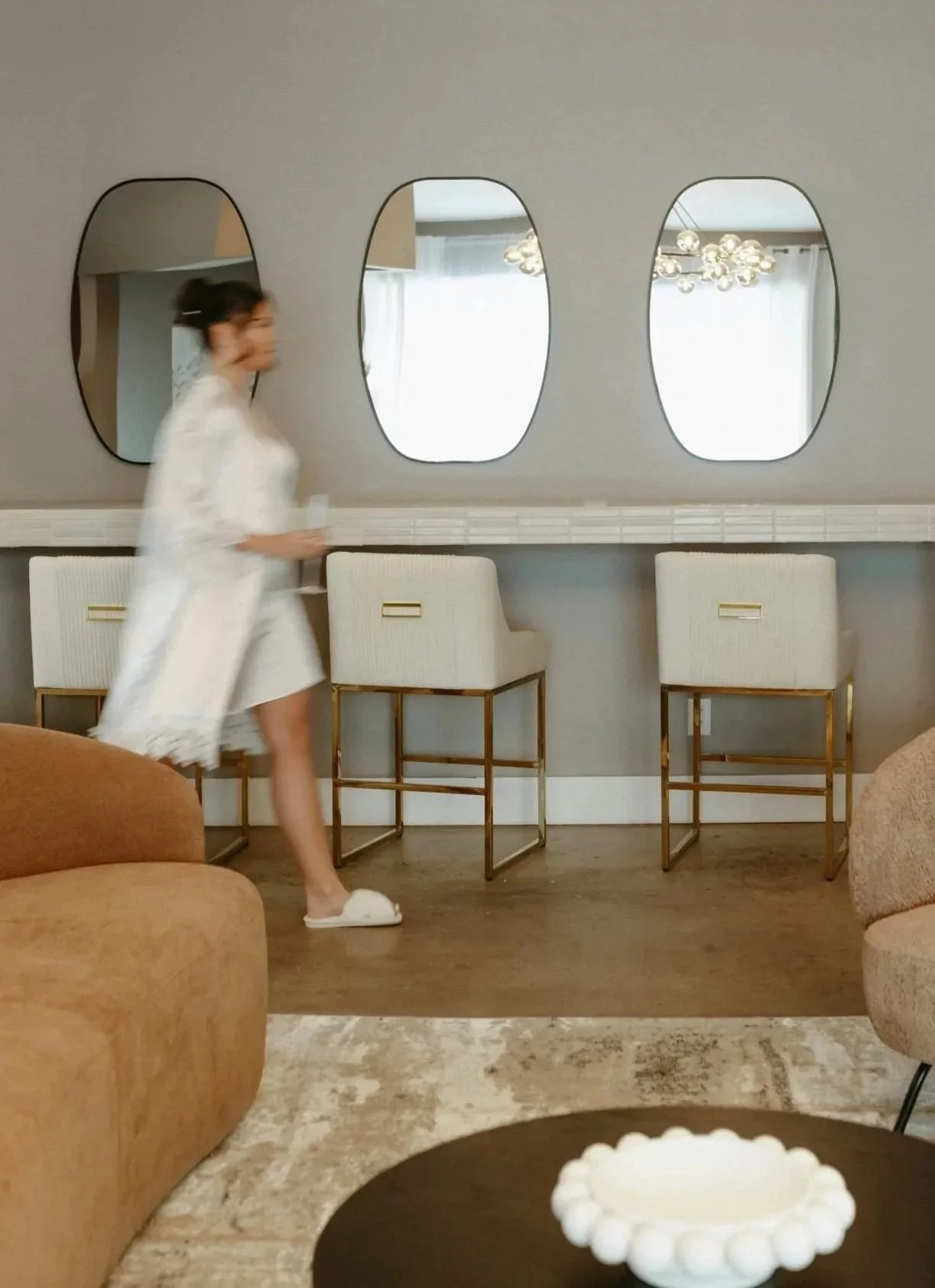 Woman in white dress walking past row of chairs and mirrors.