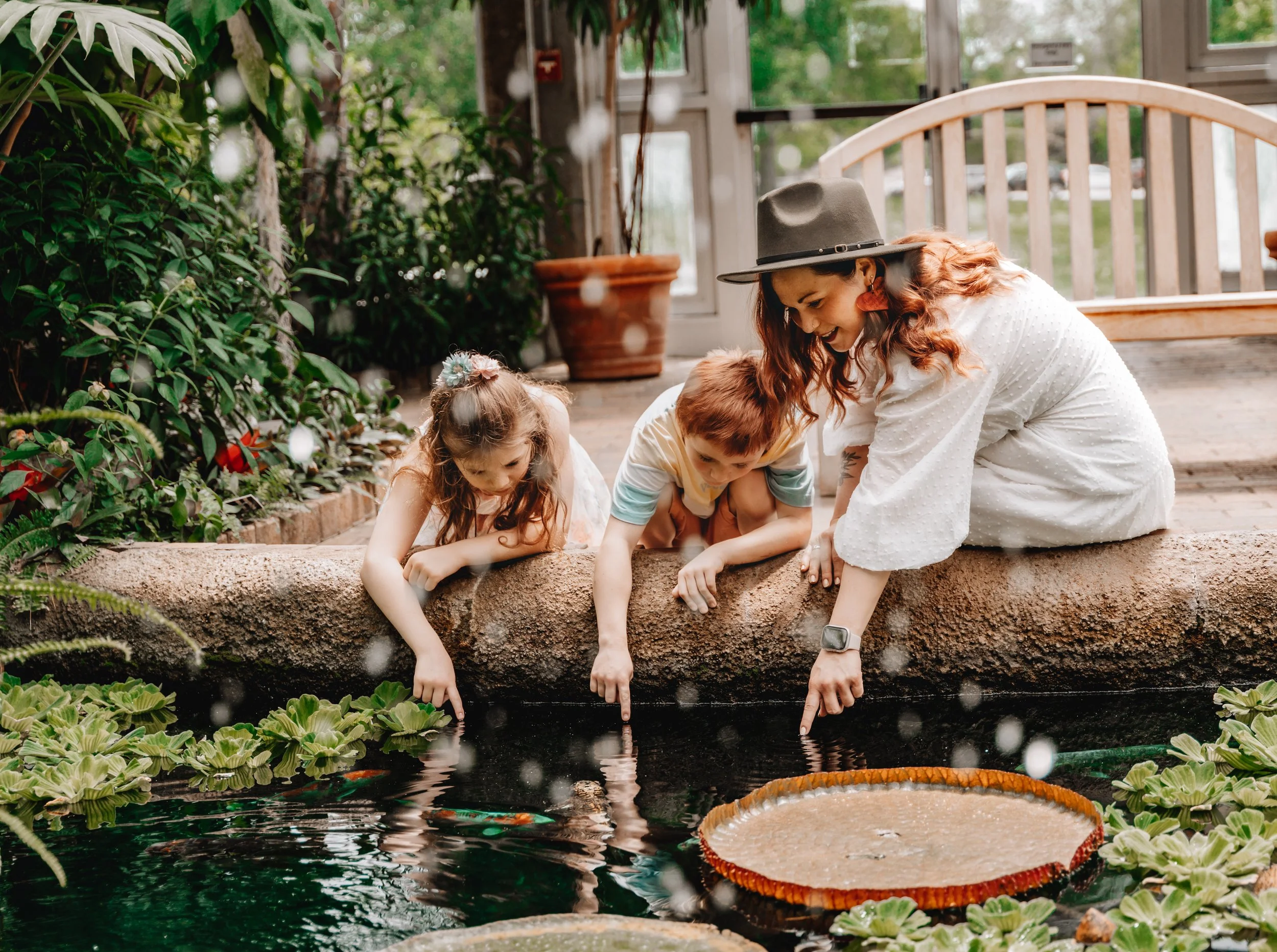 A woman and two children, a girl and a boy, lean over the edge of a pond, pointing at fish in the water. The woman wears a wide-brimmed hat, white dress, and watch, while the children have casual clothes; the girl has a flower hair accessory. They ar