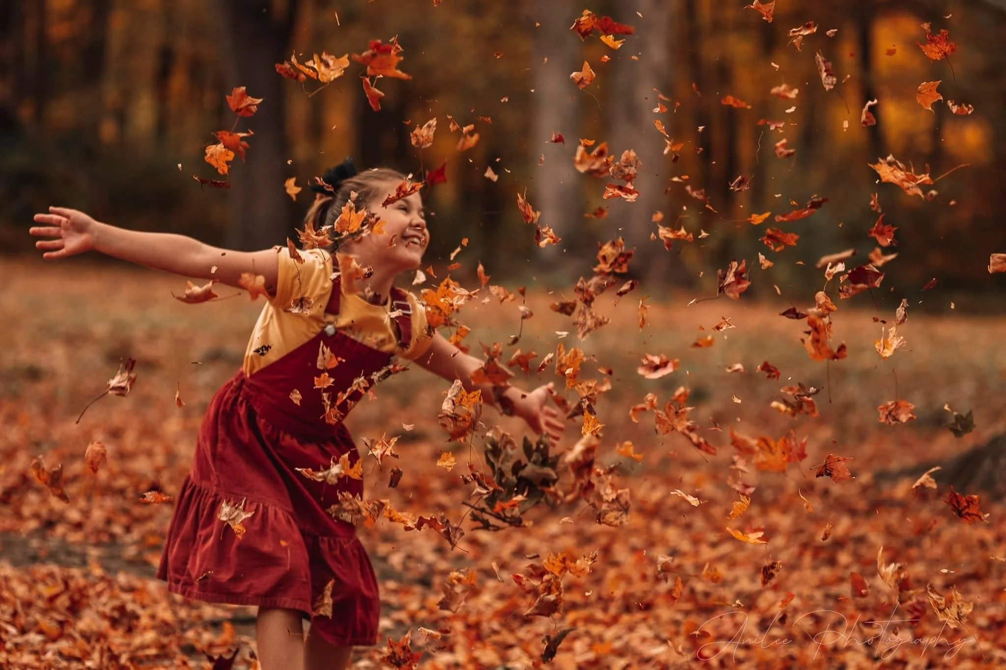 A young girl playing in a hill of fallen autumn leaves, throwing leaves into the air with a joyful expression on her face in a park with trees in the background.