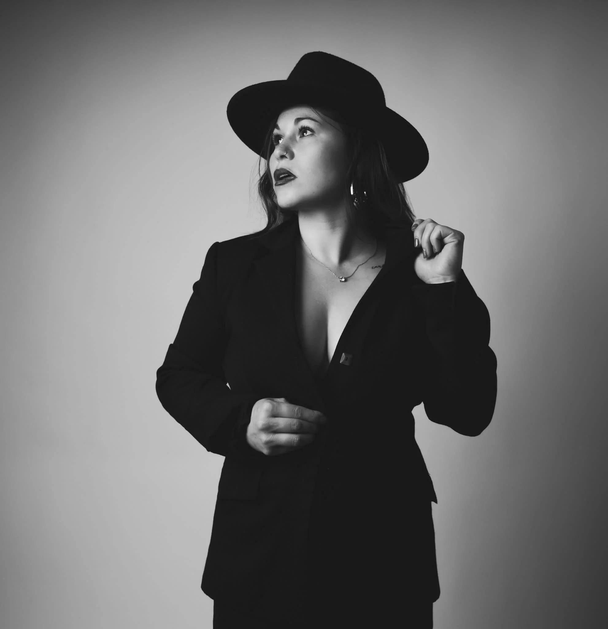 A black and white portrait of a woman wearing a wide-brimmed hat and a fitted blazer, looking to the side with a confident expression.