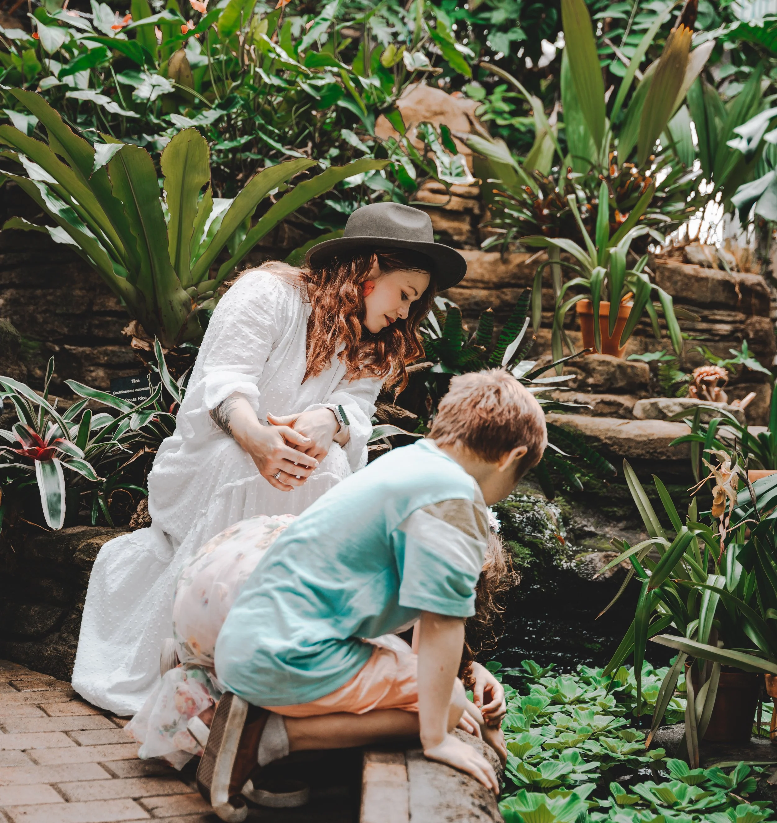 A woman with long wavy hair wearing a white dress and a gray hat, and a young boy with short hair wearing a light blue T-shirt and pink shorts, are sitting together by a lush indoor pond surrounded by green plants and rocks.
