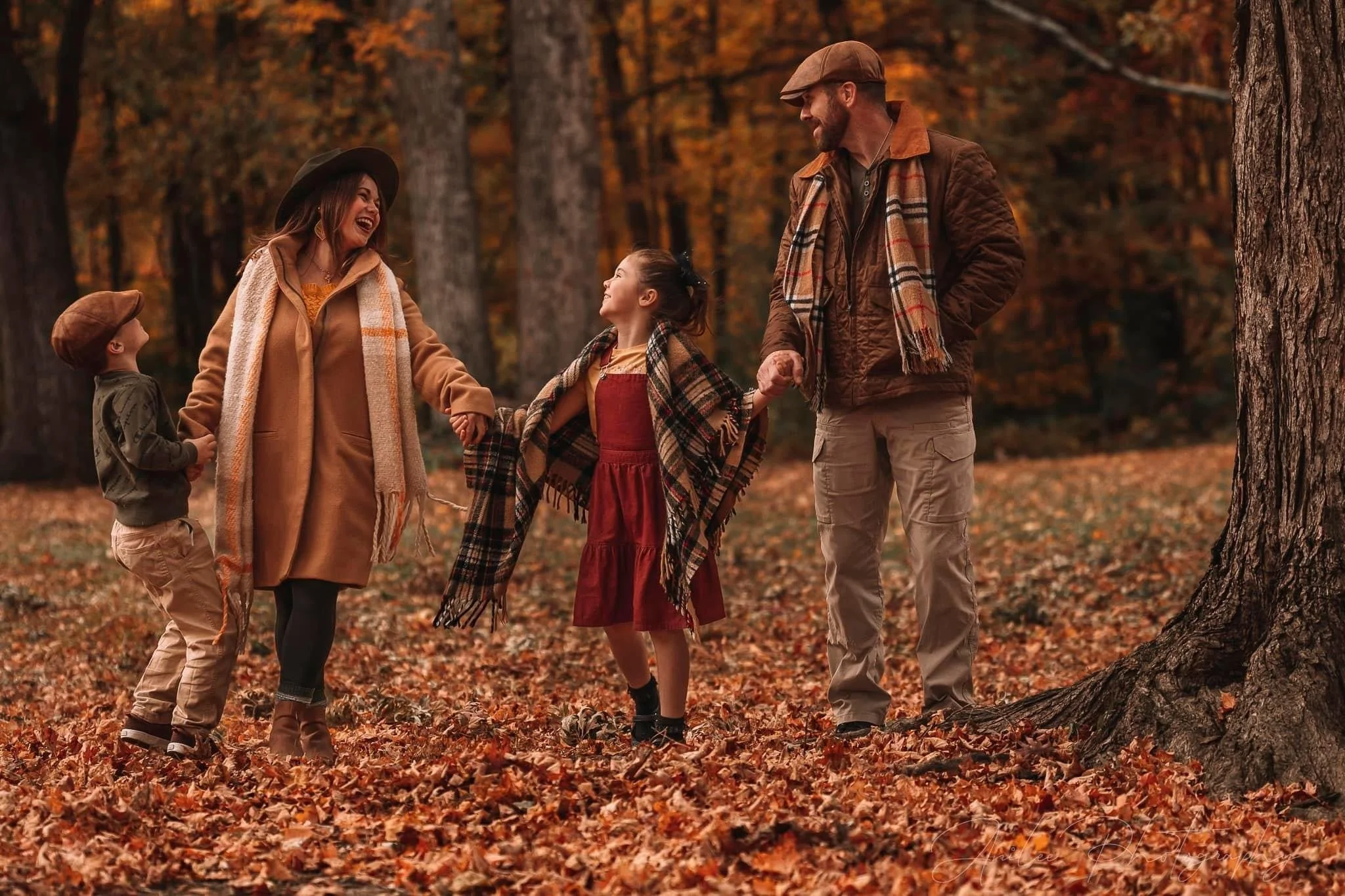 A family of four enjoying fall outdoors in a forest, holding hands and smiling. The mother and daughter are in the center, with the daughter wearing a red dress and a plaid shawl. The father is on the right, wearing a brown jacket and a plaid scarf, 