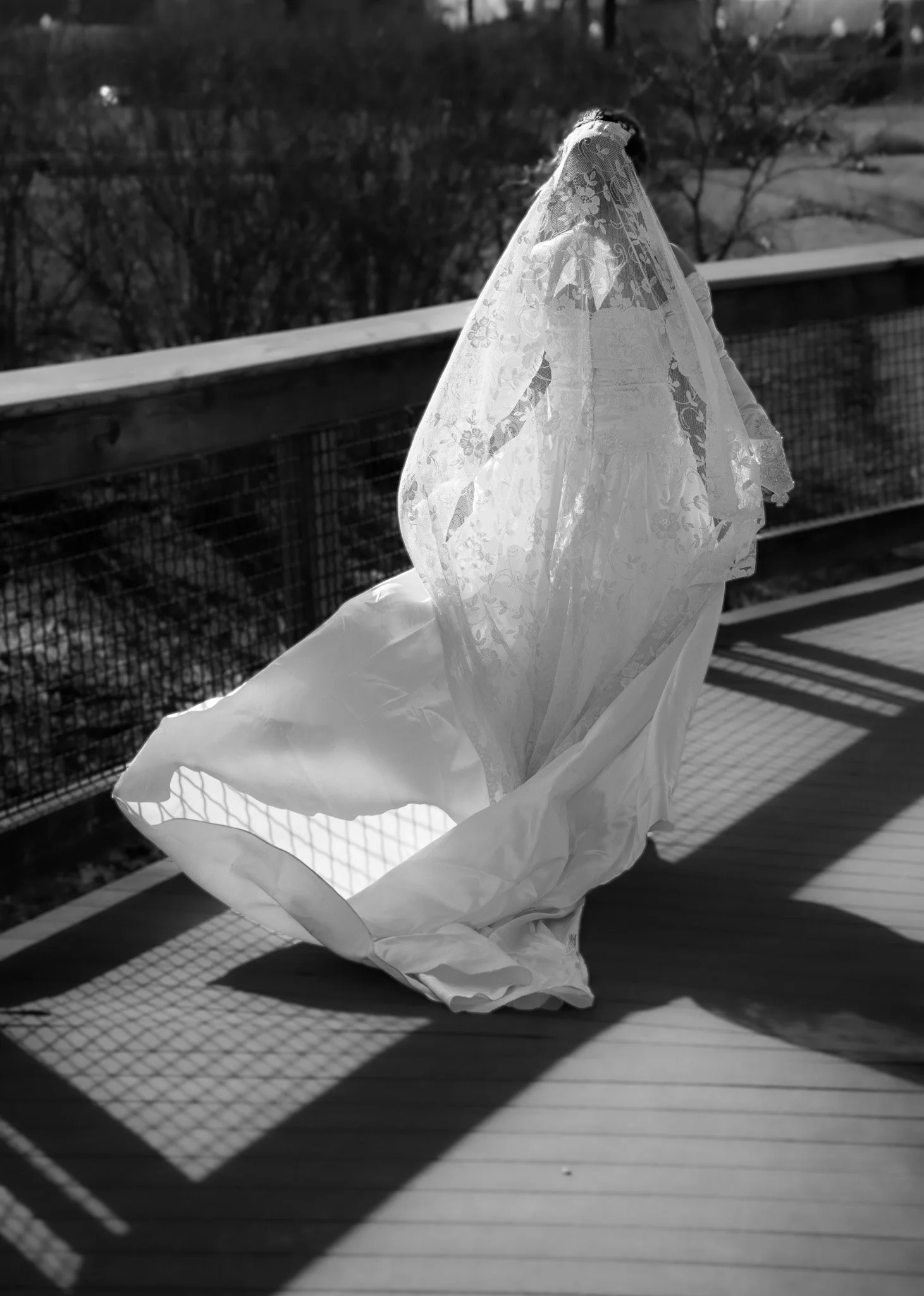 A wedding dress with a lace veil hanging on a wooden deck, with shadows cast by the railing and sunlight highlighting the fabric in black and white.