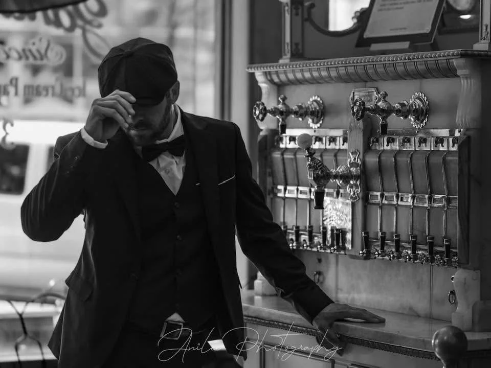 A man in a tuxedo with a bow tie, wearing a cap, standing next to a vintage soda fountain or beer-dispensing machine, adjusting his hat.