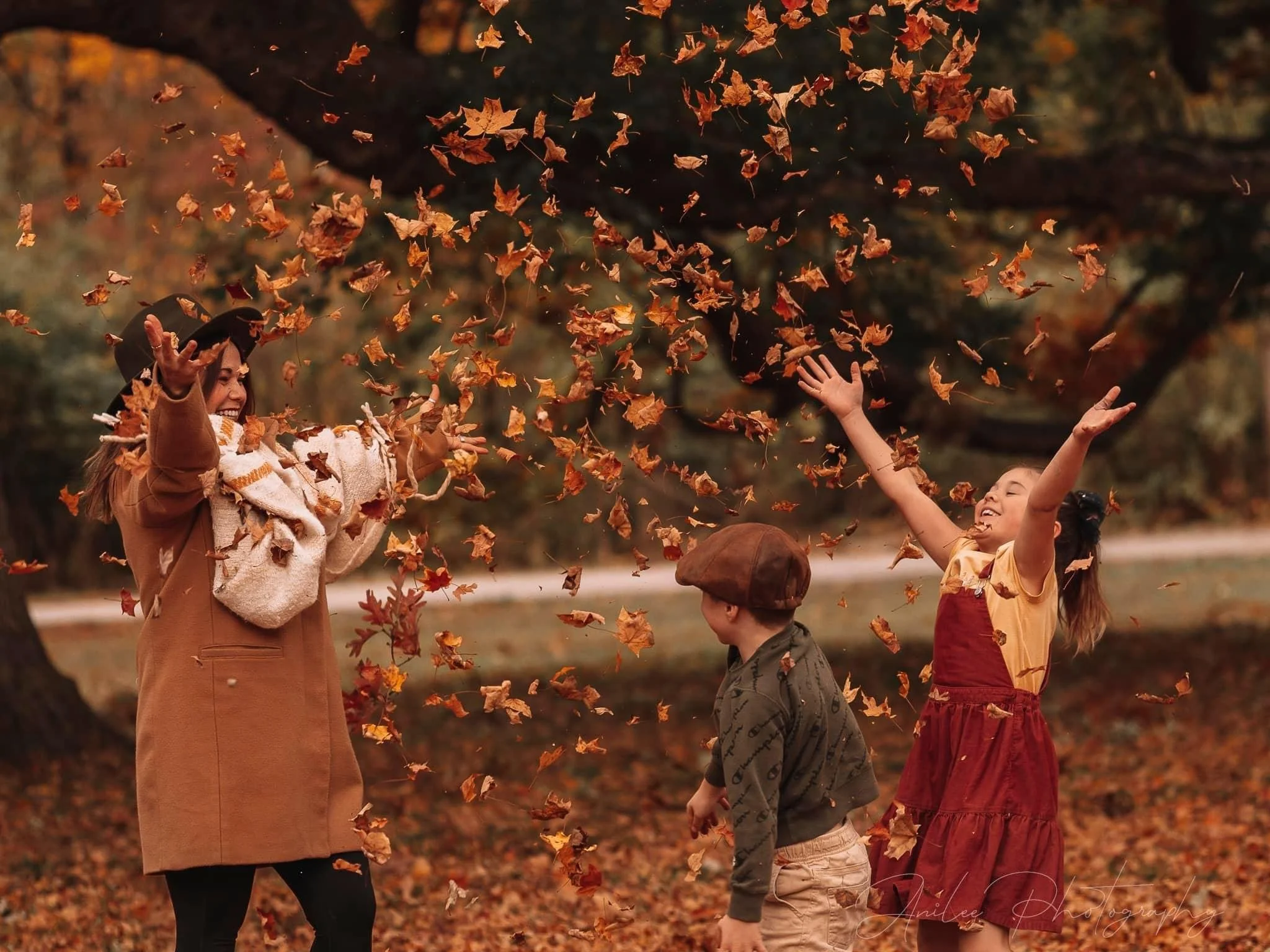 A woman and two children playing with fallen autumn leaves outdoors in a park.